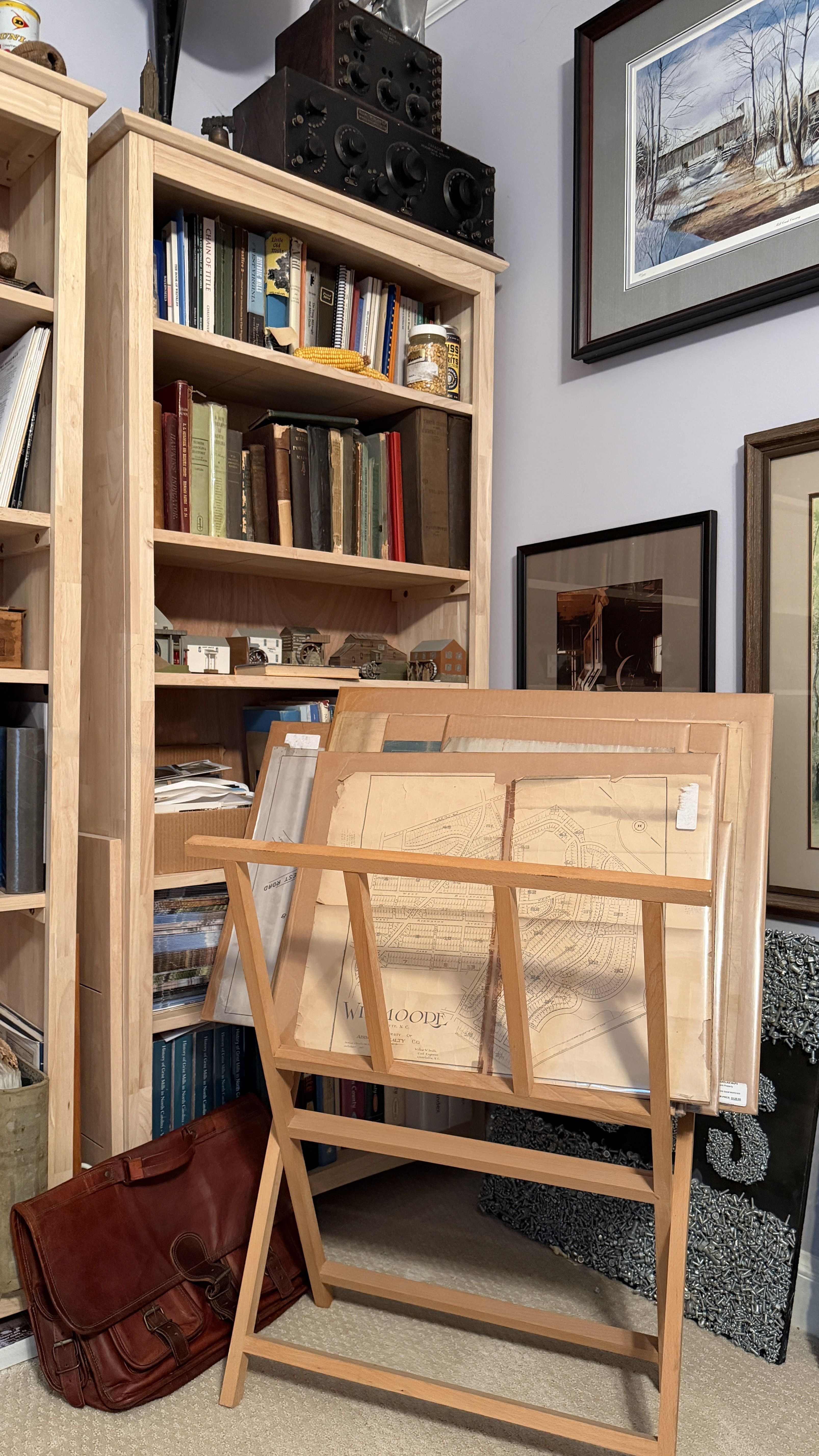 Light wood bookcase filled with books and miniature models sits beside a black electronics unit. A wooden drafting table holds large architectural drawings, with a brown leather bag on carpet.