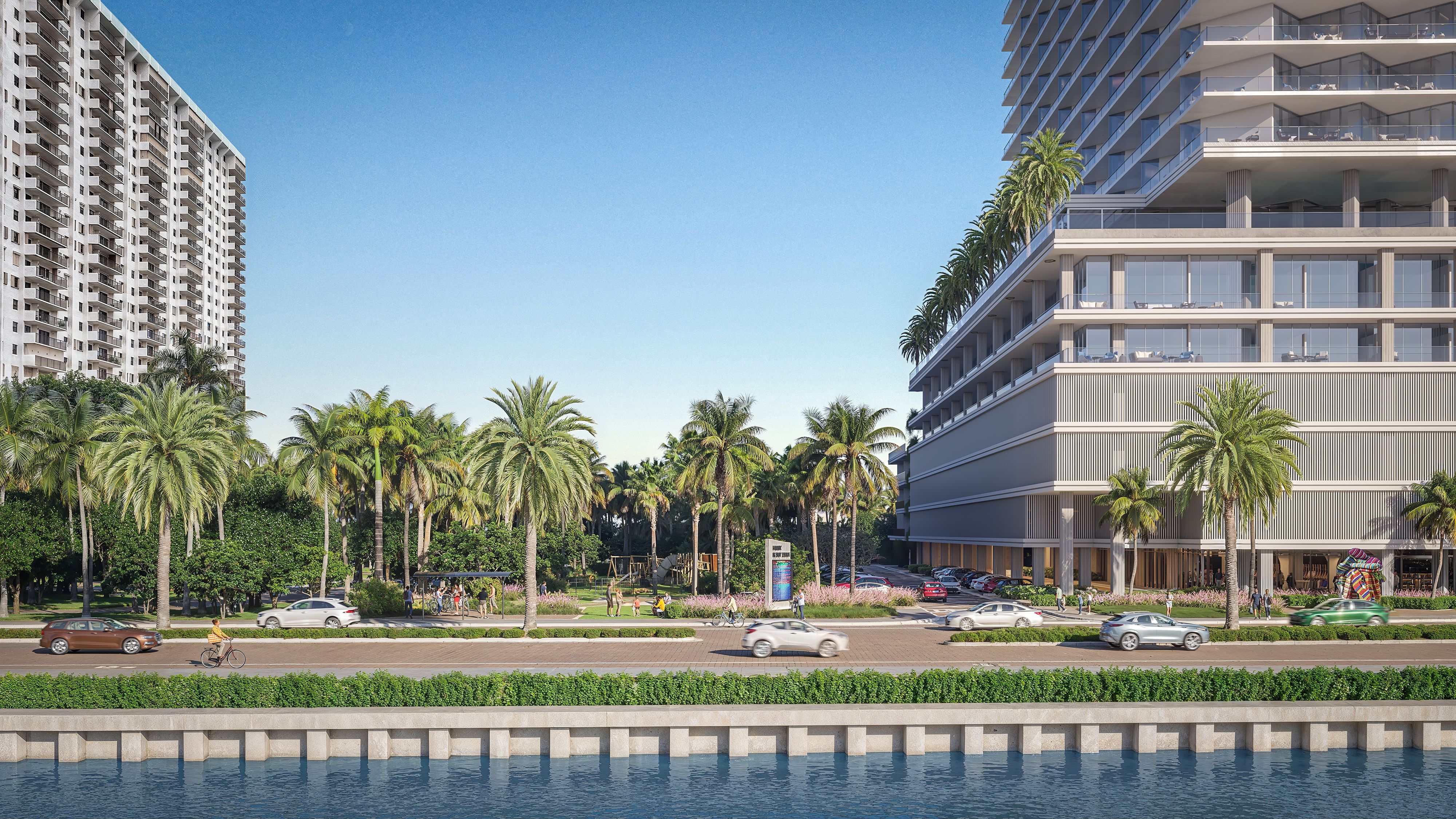 Waterfront scene with a left beige high-rise and a right glassy, tiered hotel-style building. Palm trees line a sunny park as pedestrians and cyclists pass; cars drive along the street by the calm blue water.