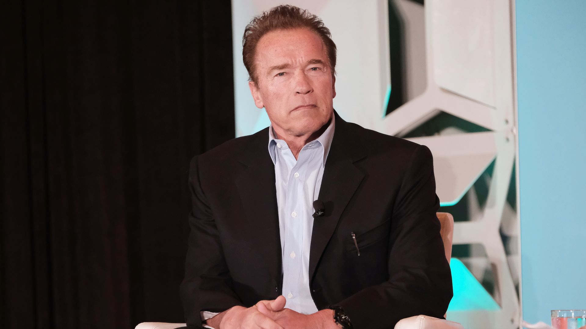 Arnold Schwarzenegger sitting down in a while chair with a serious expression
