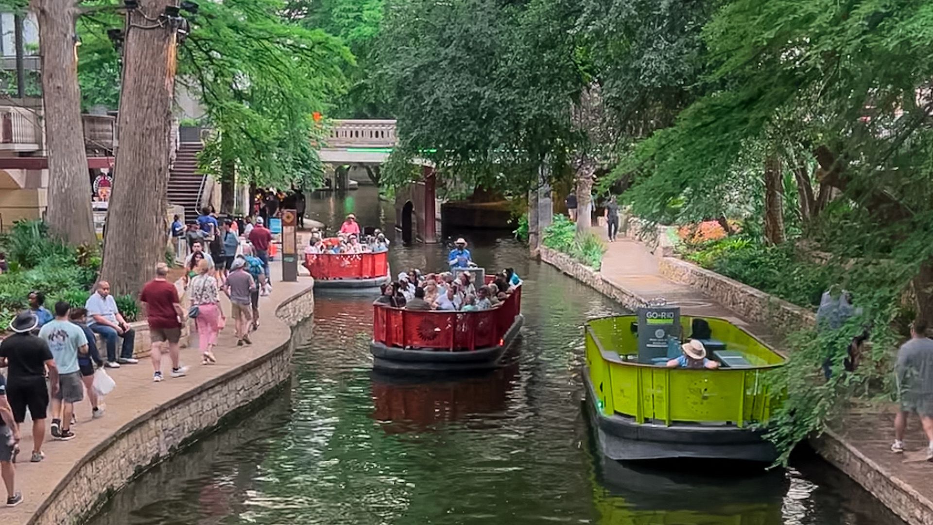 People stroll along the San Antonio River Walk as colorful Go Rio barge tours glide through the water, surrounded by lush trees and historic architecture.