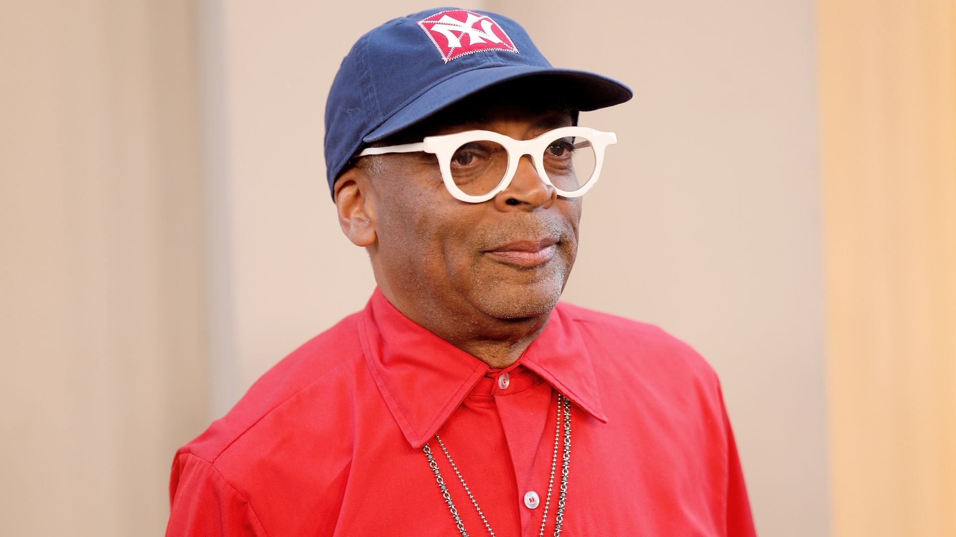 A man wears white-rimmed round glasses, navy blue hat with the New York Yankees logo affixed on a red square, red collared and buttoned shirt and necklaces. The background is beige and tan.
