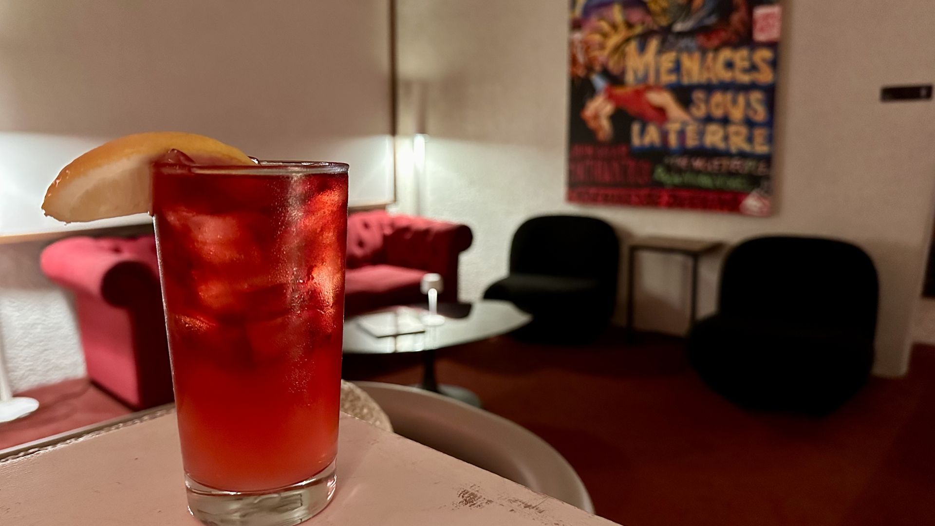 A photo of a tall reddish drink on a platform in front of a lounge with a movie poster hanging on the wall 