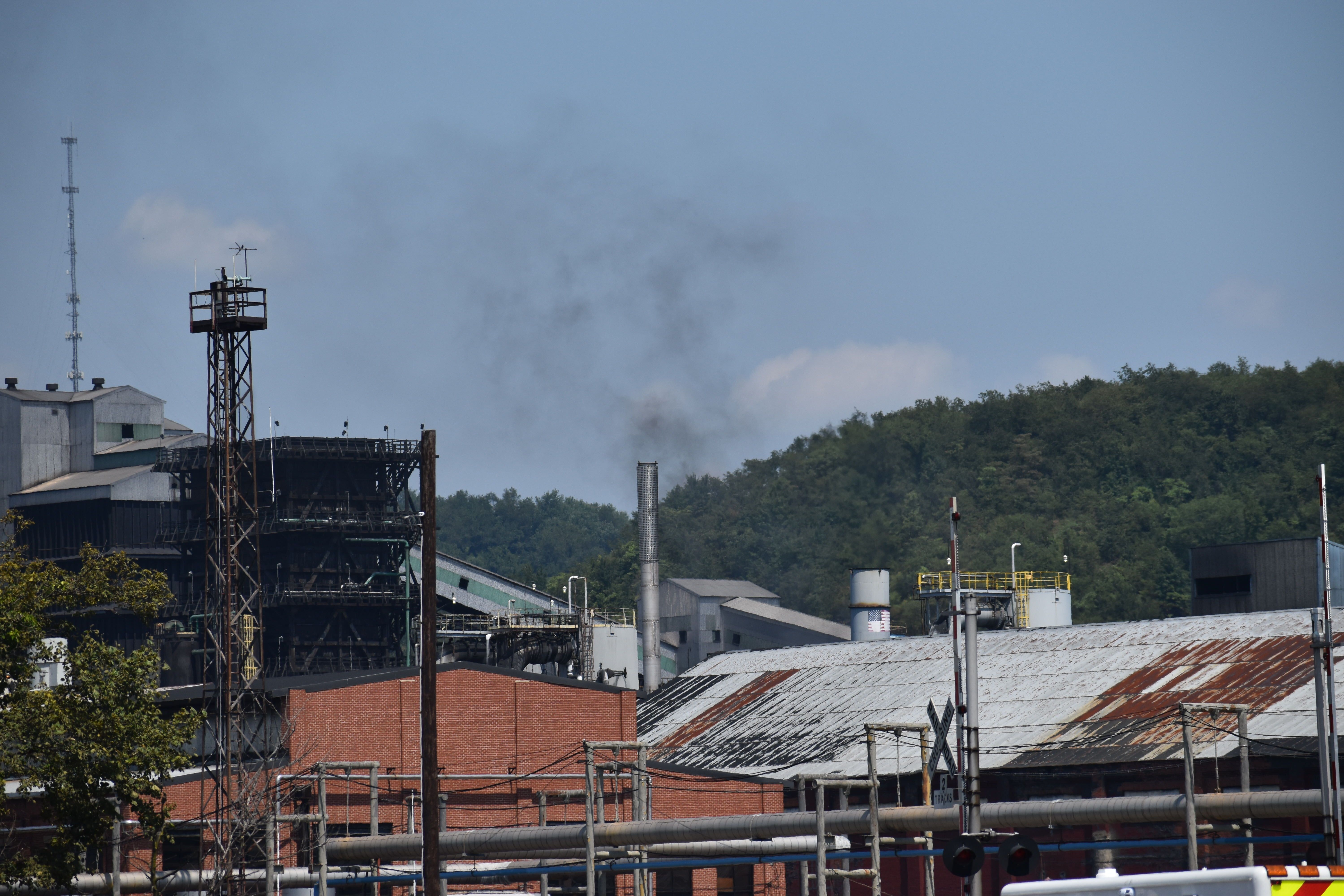 Industrial factory buildings with rusted roofs and a chimney emitting black smoke against a blue sky with green forested hills in the background.