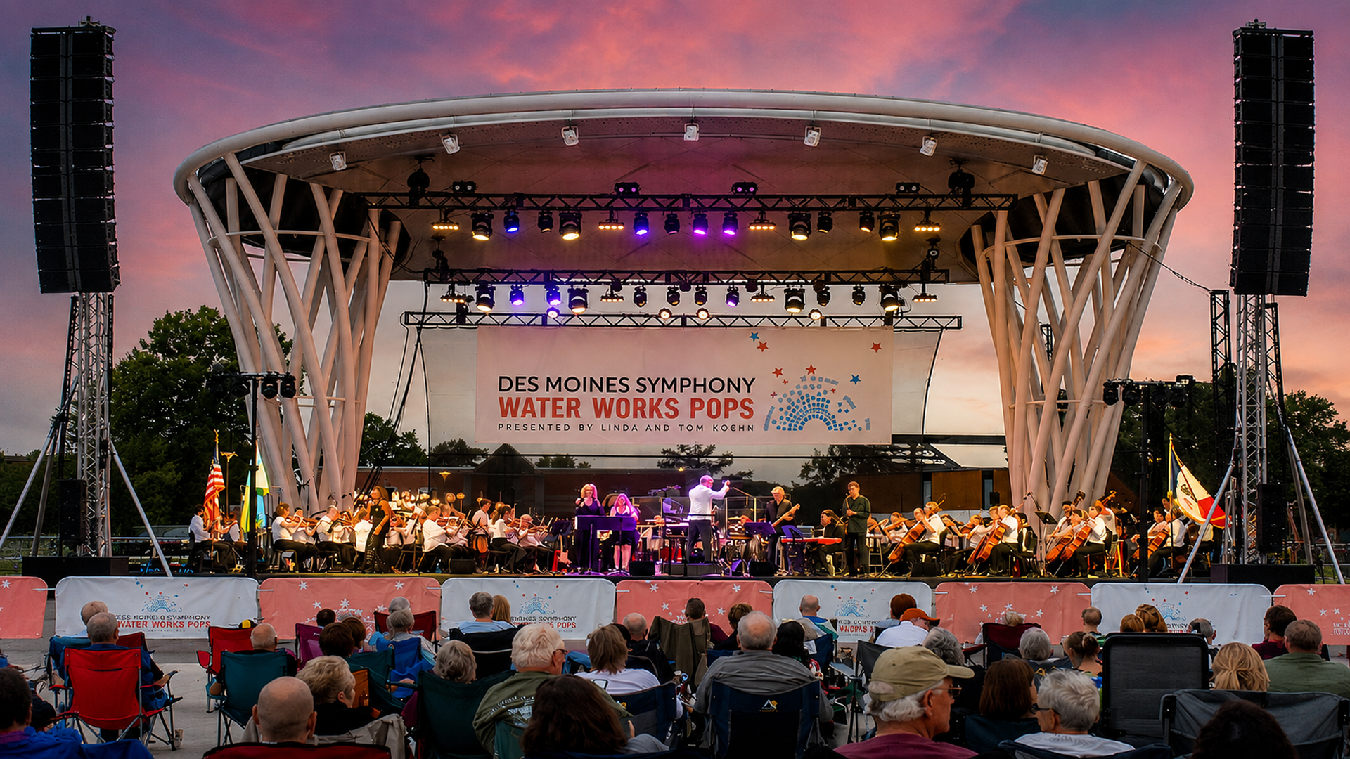 Outdoor concert stage at sunset with a full orchestra under a curved white pavilion. A banner reads "Des Moines Symphony Water Works Pops." Audience in folding chairs.