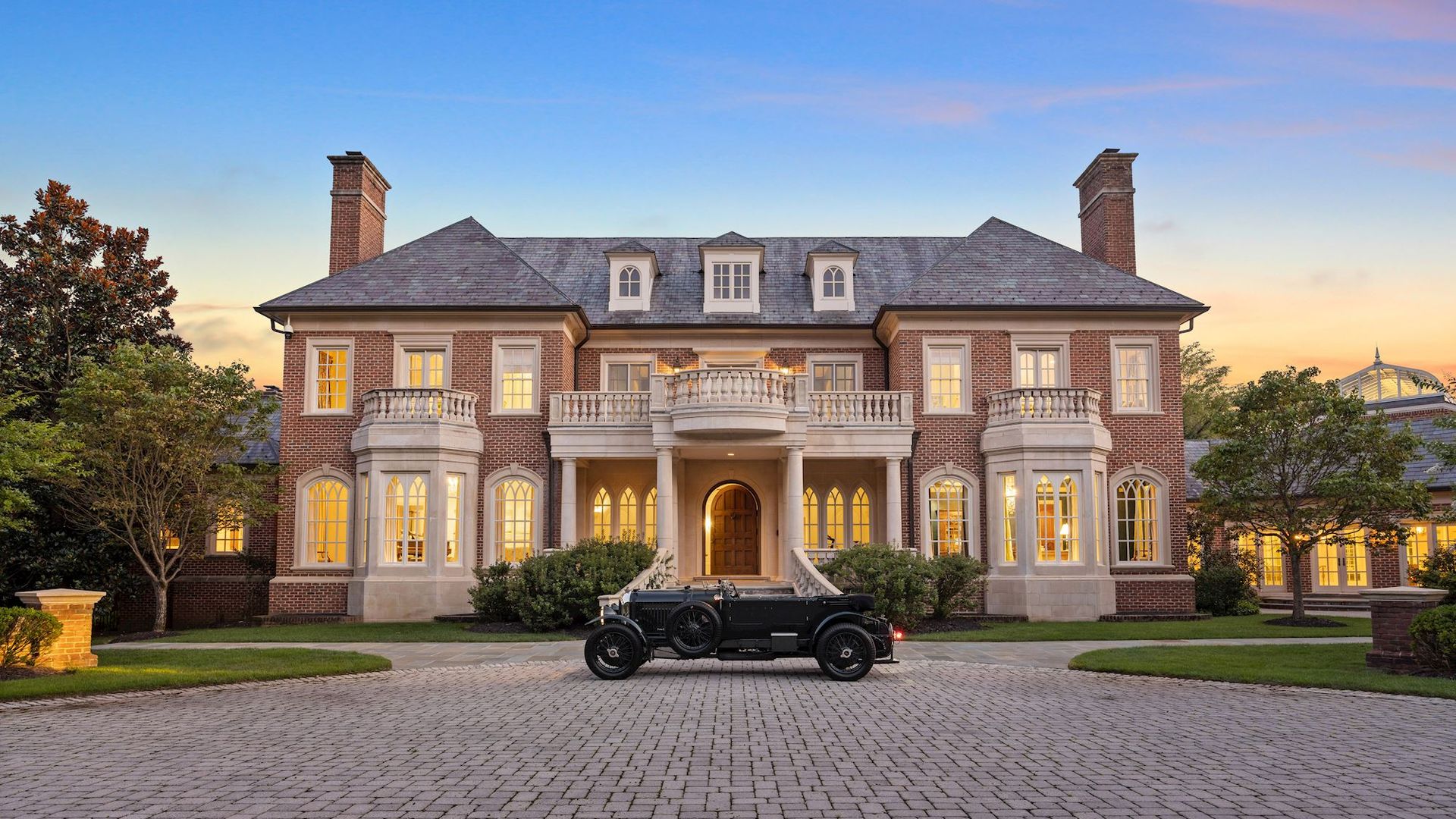 The exterior of a brick mansion with a wrap-around driveway.