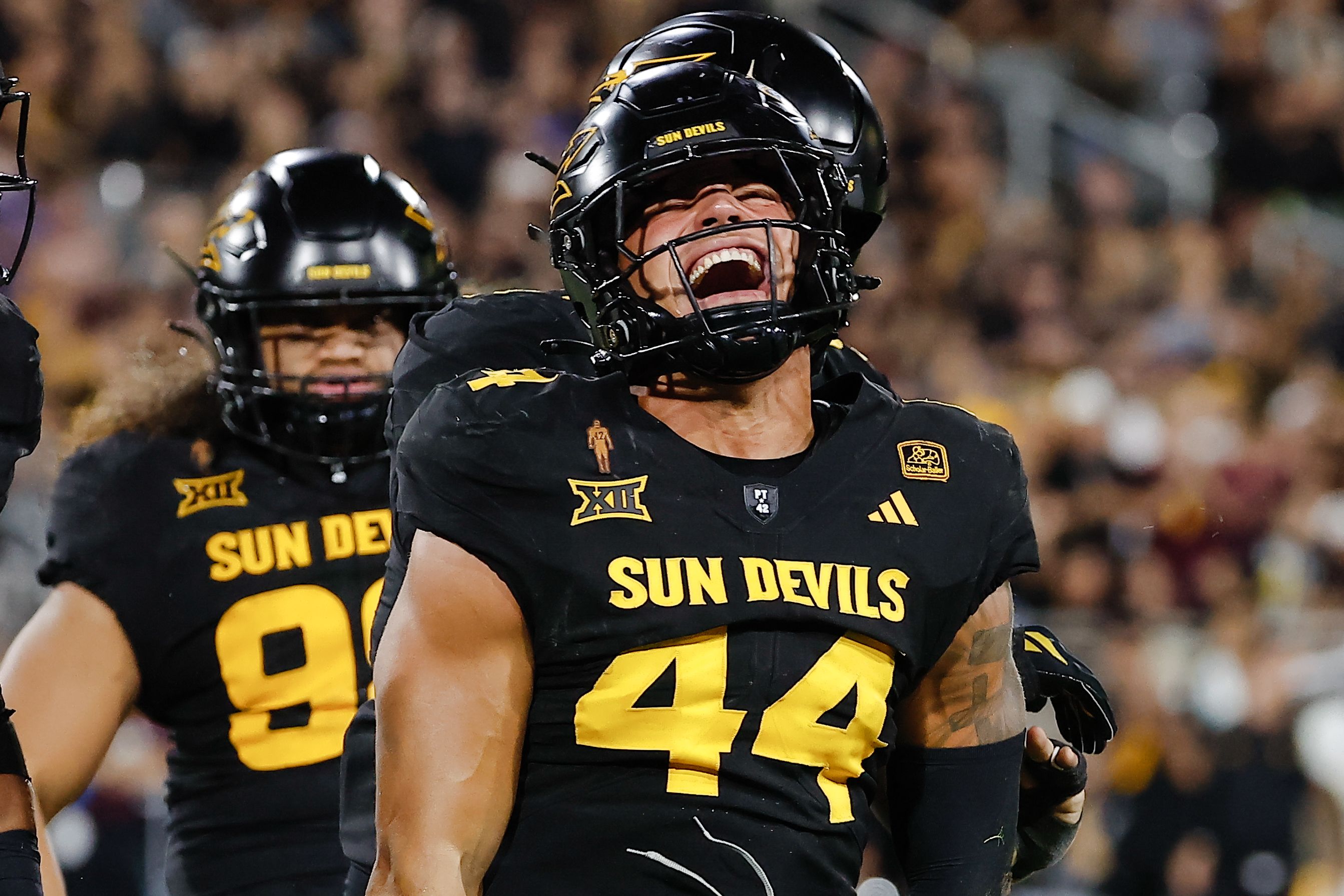 Sun Devils football players in black and gold celebrate on the field; central player grins with mouth open under a helmet, teammates nearby, blurred stadium crowd behind them.