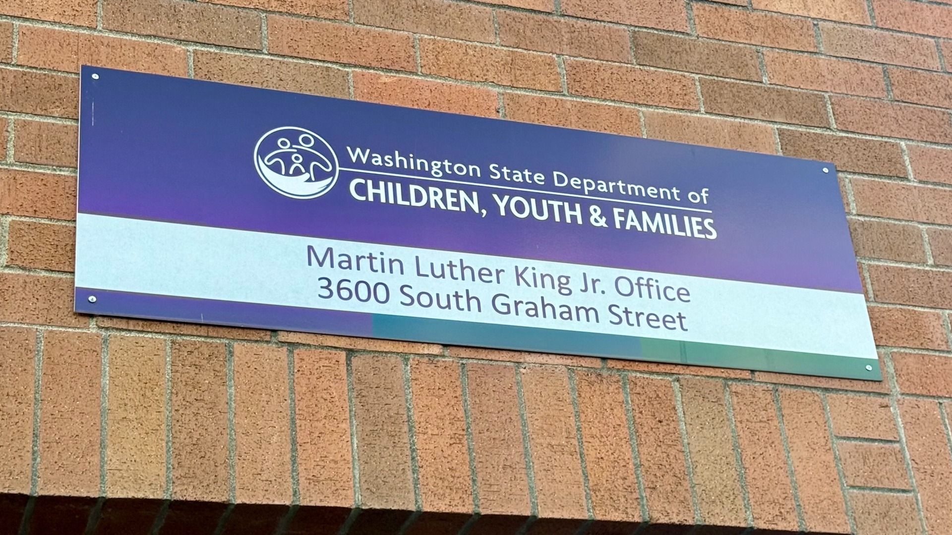 Sign on a brick wall for Washington State Department of Children, Youth & Families. Purple gradient panel with white text and circular logo; lower band shows MLK Jr. Office and 3600 S. Graham St.