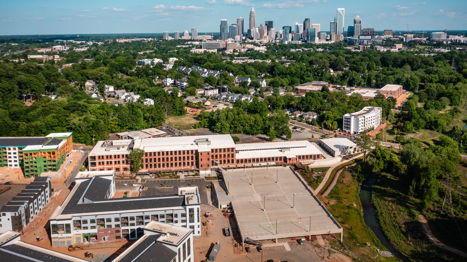 Aerial view of a city skyline with tall skyscrapers on the horizon, surrounded by green trees and suburban blocks. Foreground shows red-brick buildings, a large parking lot, and a river nearby.