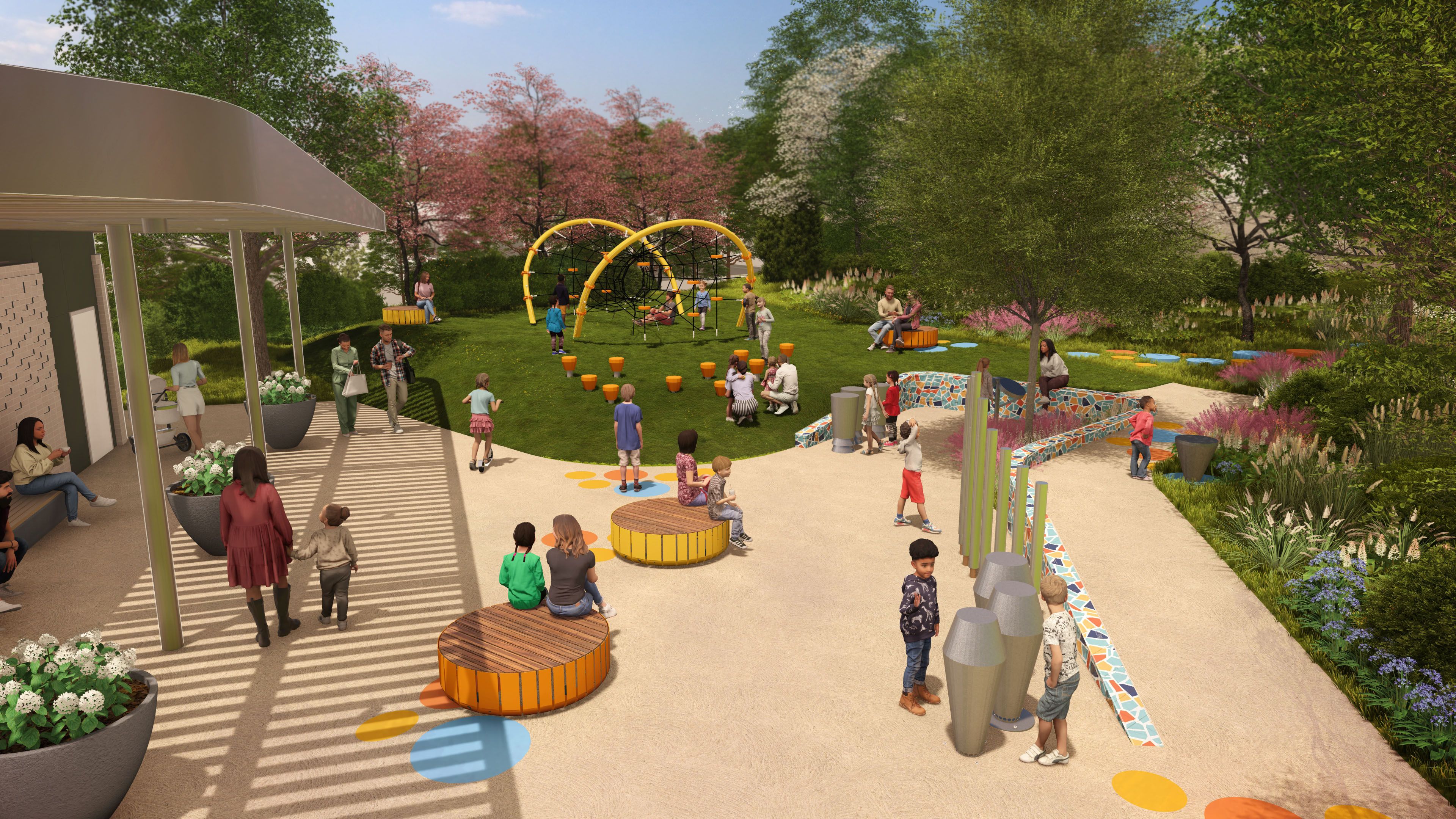 Sunny park playground filled with people. Bright yellow climbing rings, orange stools, and circular wooden benches amid green trees, flowering shrubs, and a modern building on the left.