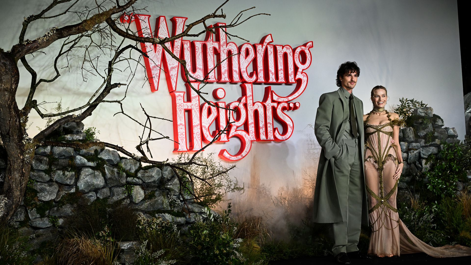 Actors Jacob Elordi, in a grey suit and long jacket,  and Margot Robbie, in a semi-sheer pale pink gown, stand arm in arm in front of a backdrop with the text "Wuthering Heights."