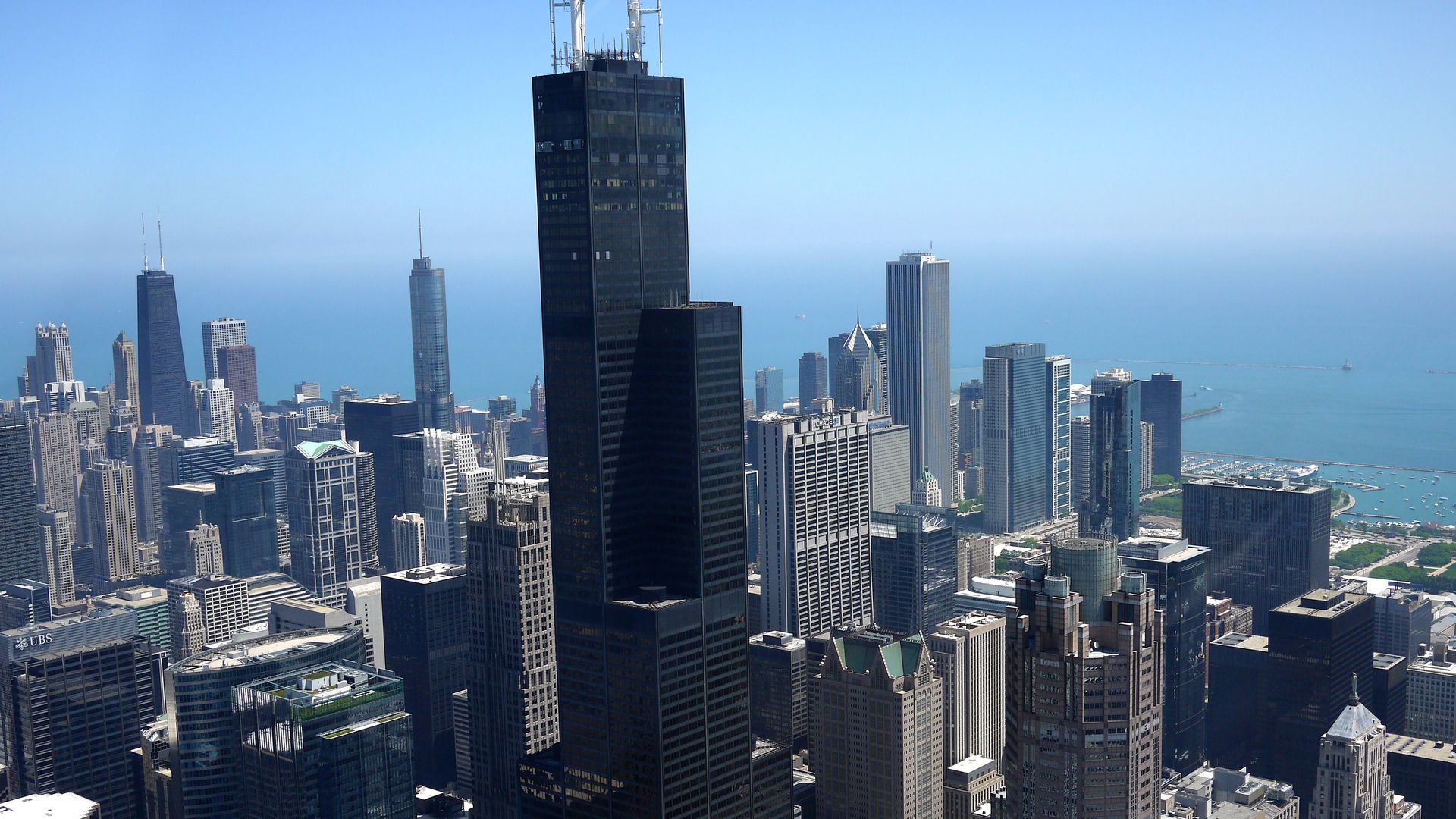 Aerial view of a dense downtown skyline with the tall, dark Willis Tower at center, surrounded by glass skyscrapers, and a blue Lake Michigan horizon in the distance.