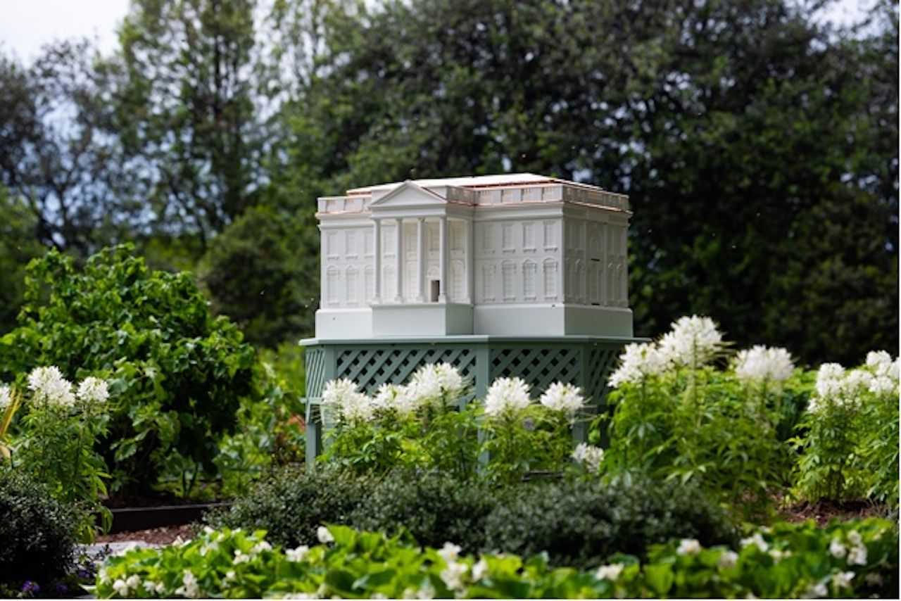 White neoclassical miniature building on a green lattice pedestal sits in a garden with white flowering plants and dense greenery, trees in the background.
