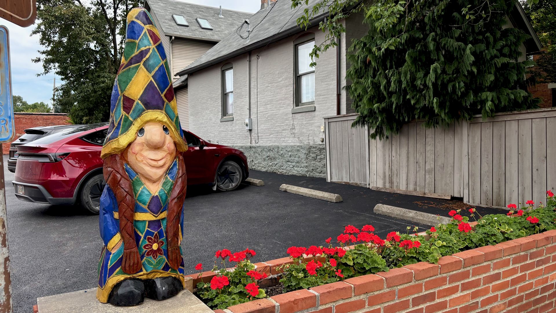 Colorful, patchwork gnome statue in a parking lot beside a red car. A gray brick house, wooden fence, and red flowers along a brick planter frame the scene.