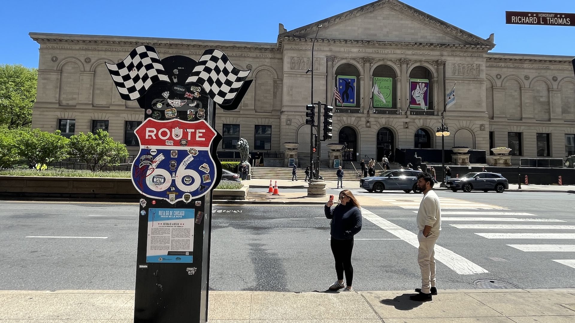 Sunny street scene in front of the Art Institute of Chicago’s neoclassical façade, with a large Route 66 sign and black-and-white checkered flags, pedestrians, and a clear blue sky.
