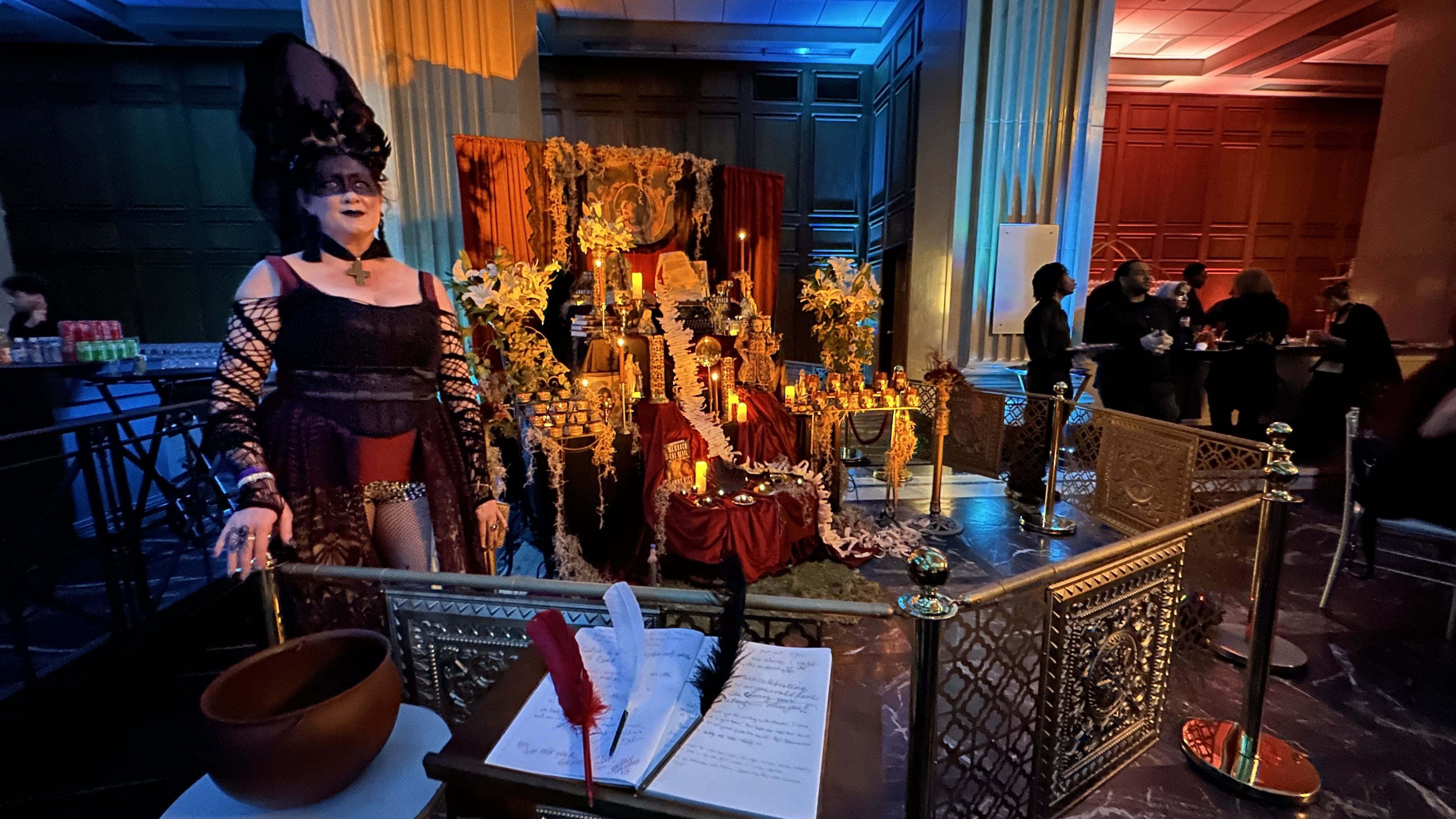 Woman in black gothic costume with tall headpiece stands near a red and gold altar decorated with candles, flowers, and skulls in a dimly lit room with people and blue-orange lights.