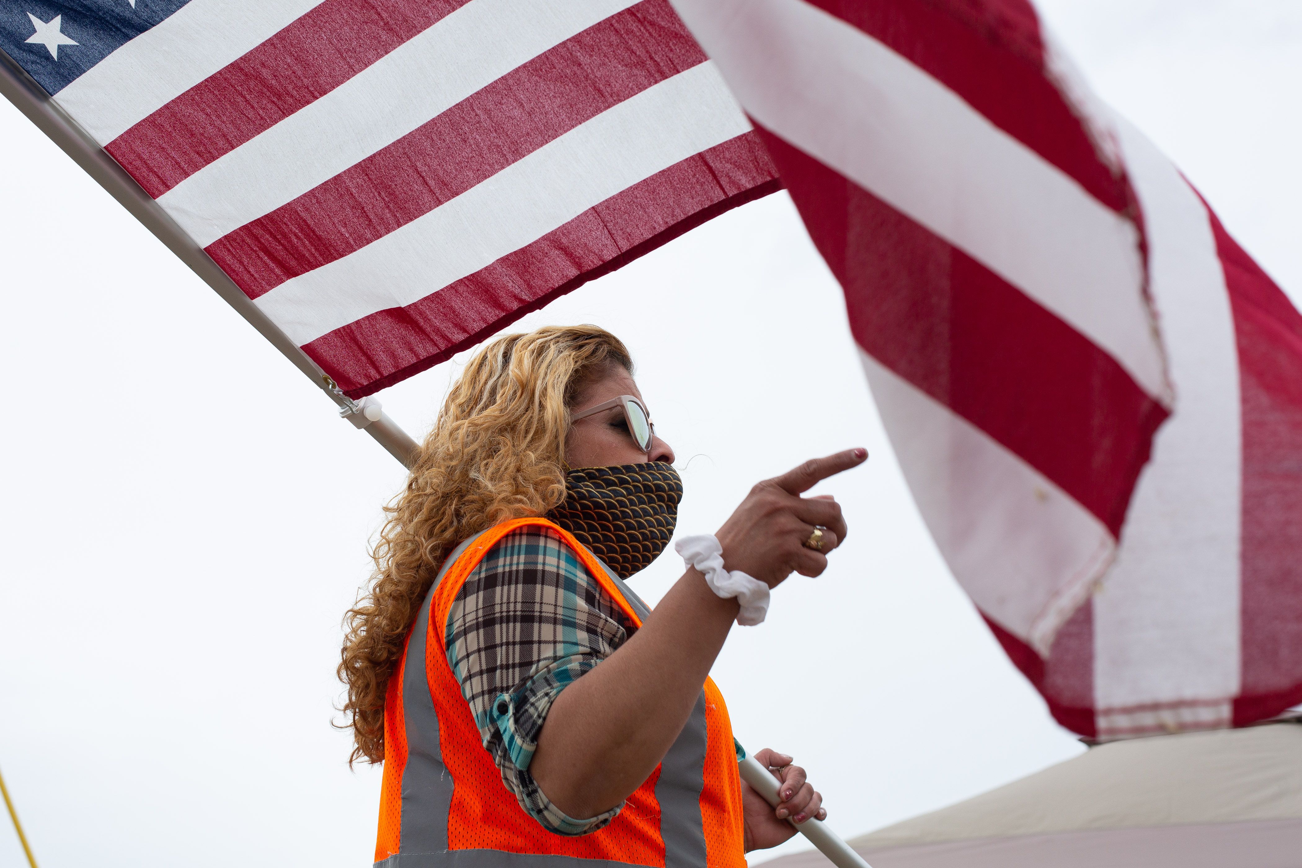 A person wearing a safety vest and face mask points a finger while holding an American flag.