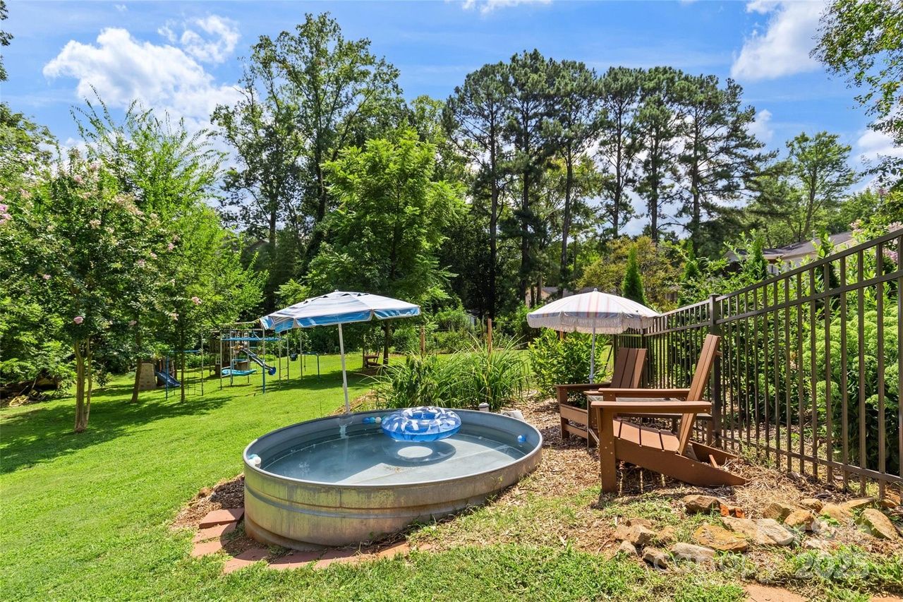 Outdoor backyard scene with a round metal water tub, blue inflatable ring, two brown wooden chairs under striped umbrellas, surrounded by green grass, trees, and a metal fence.