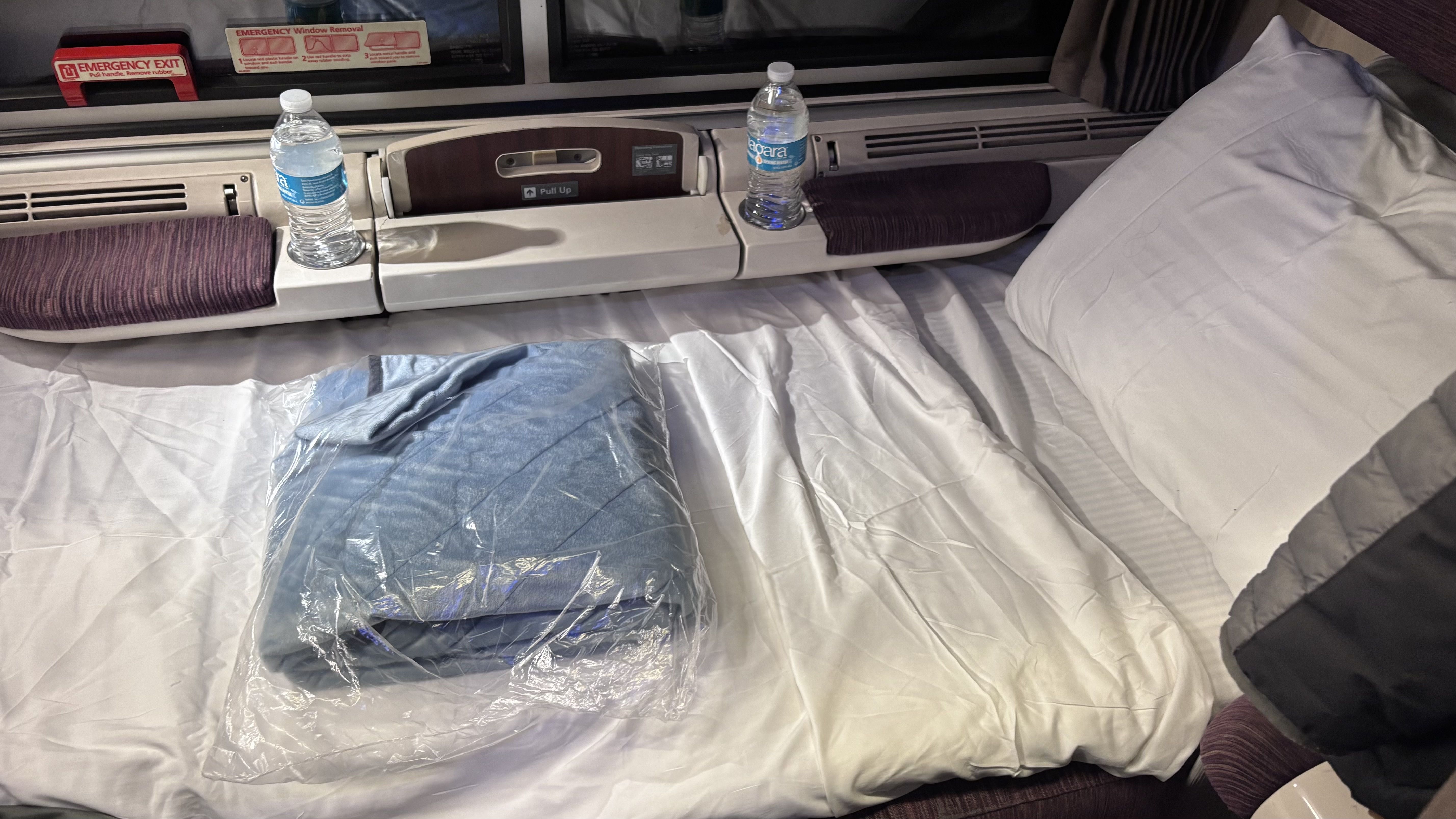 A compact bed on a train or bus with white sheets, a pillow, two bottles of water on a shelf, and a blue blanket wrapped in plastic on the bed.