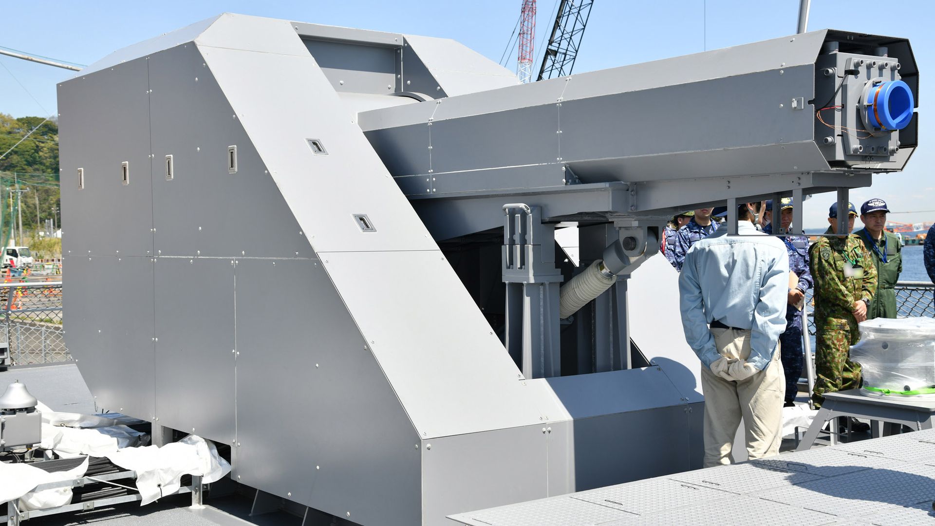A closely cropped photo of a railgun aboard a Japanese naval ship. People in military uniform are seen in the background. Cranes loom overhead.
