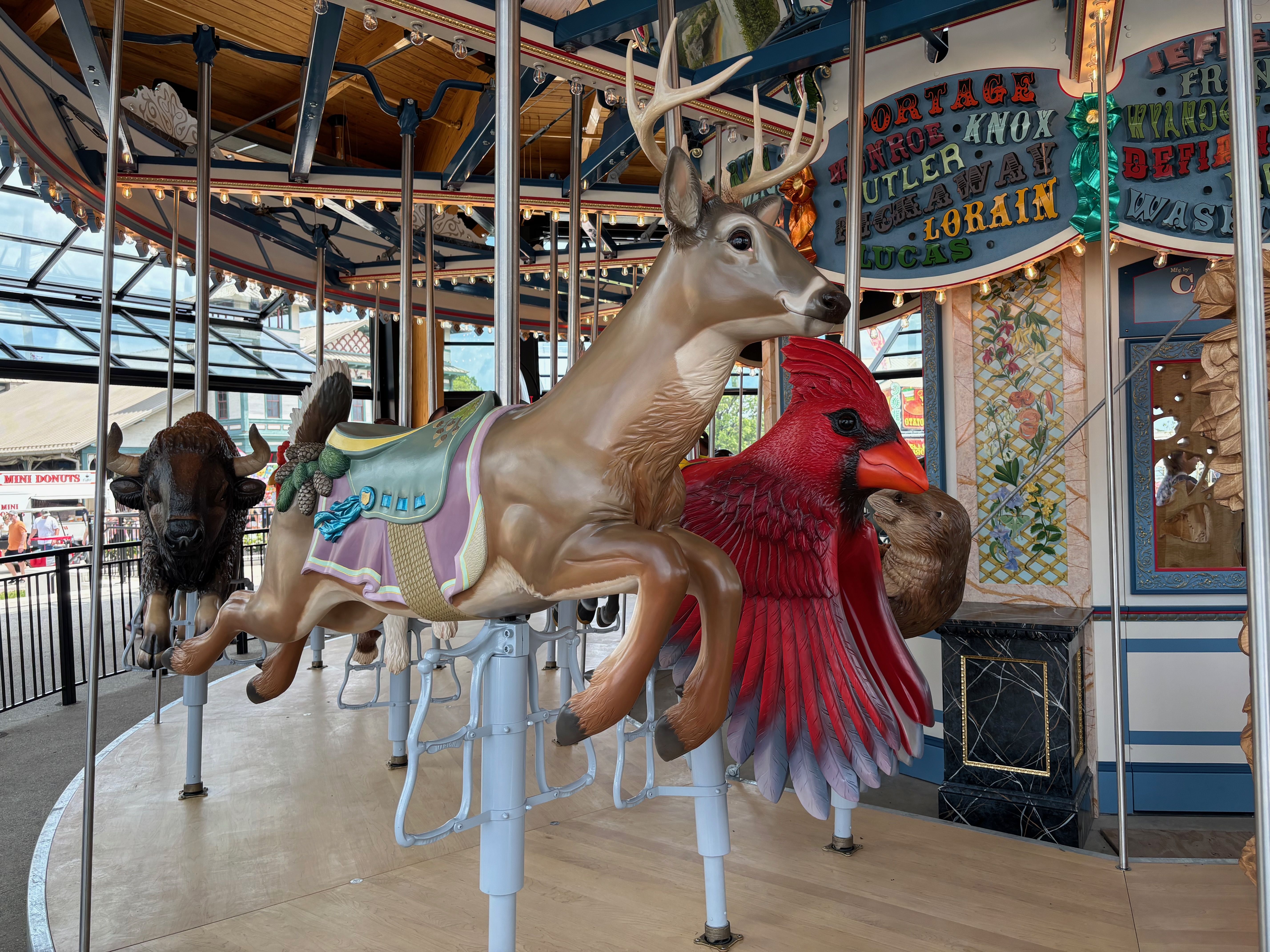Indoor carousel with ornate animal figures; a beige deer with antlers beside a red cardinal, both mounted on poles. Bright string lights, blue trim, and decorative paneling in the background.