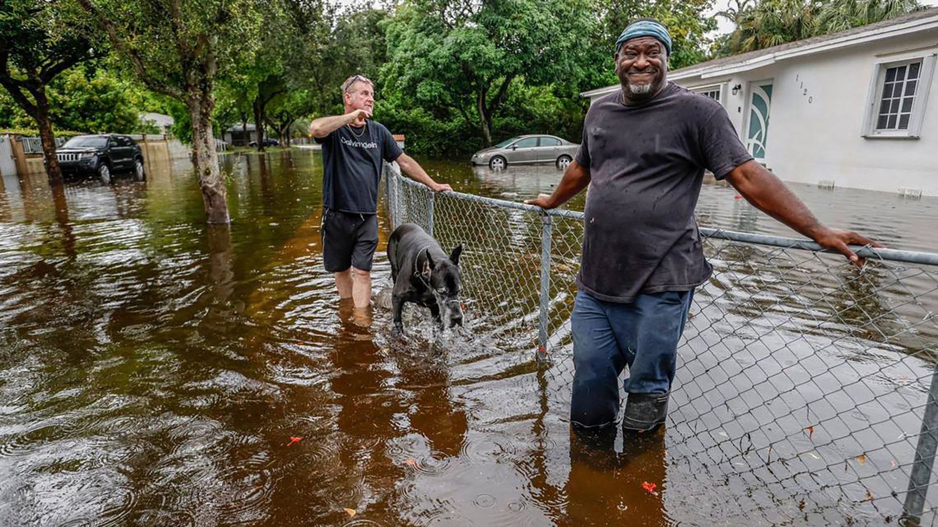 Two men and a black dog stand in knee-deep floodwater outside a house; one man leans on a chain-link fence, a car and trees partly submerged in water around them.