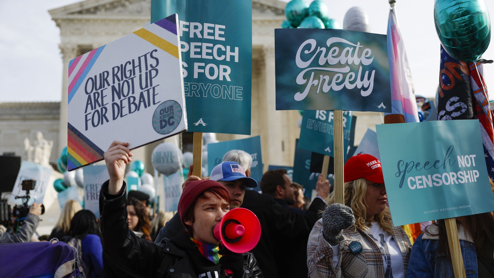A person with a red megaphone holds up a sign with the words OUR RIGHTS ARE NOT UP FOR DEBATE while surrounded by other people holding up signs outside a classical building.