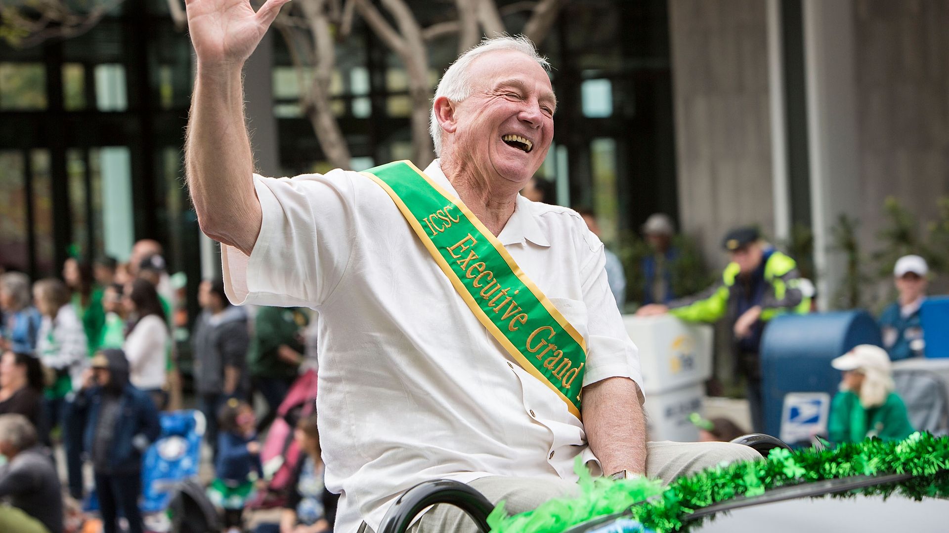 Jerry Sanders wearing a green sash and riding in a convertible car waiving to the crowd as the honorary grand marshal at a St. Patrick's Day parade through Balboa Park
