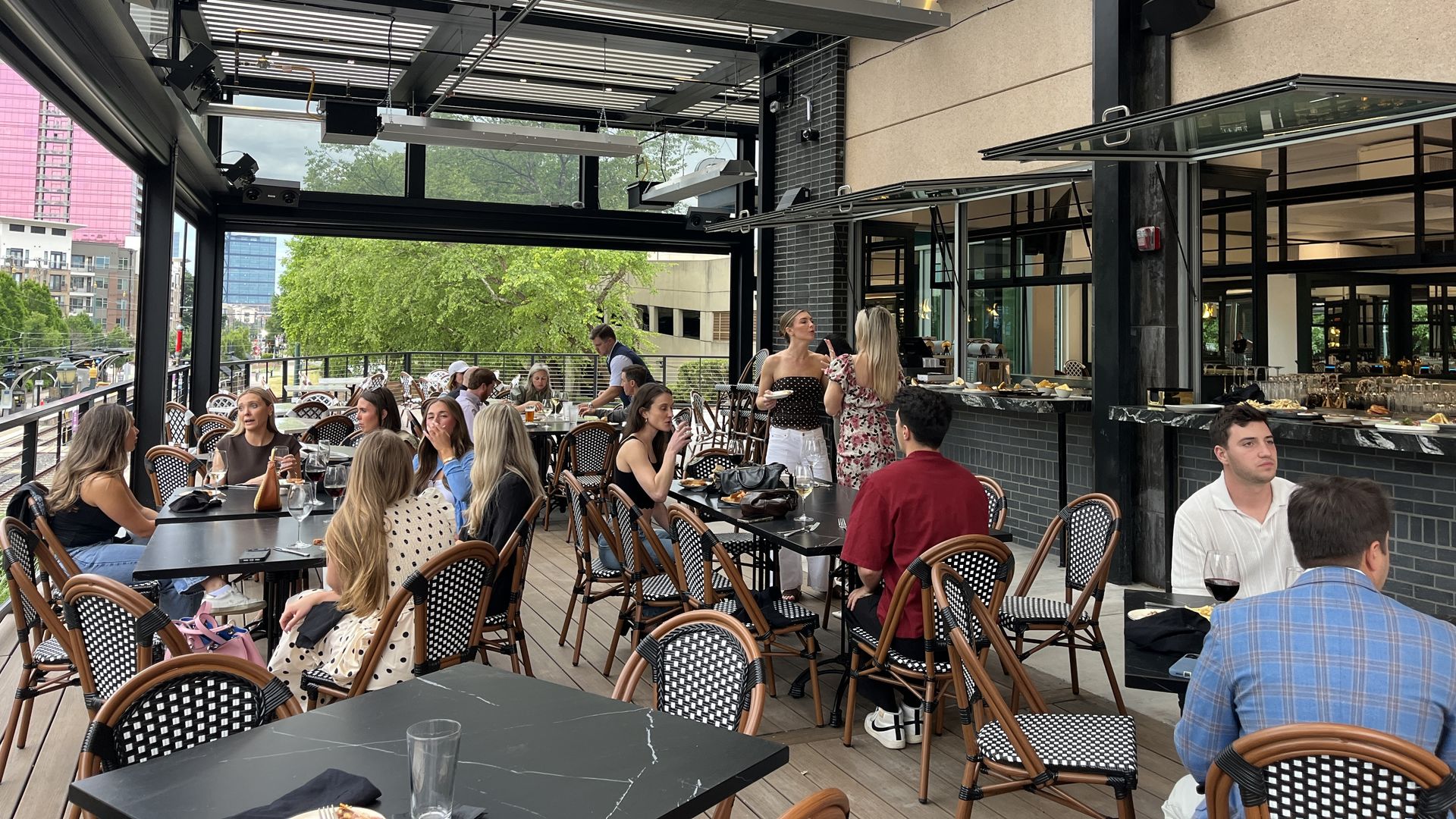 Outdoor restaurant terrace crowded with diners at black marble tables and wicker chairs. Waitstaff mingle near a serving counter under a modern industrial ceiling; glass walls reveal trees and distant buildings.