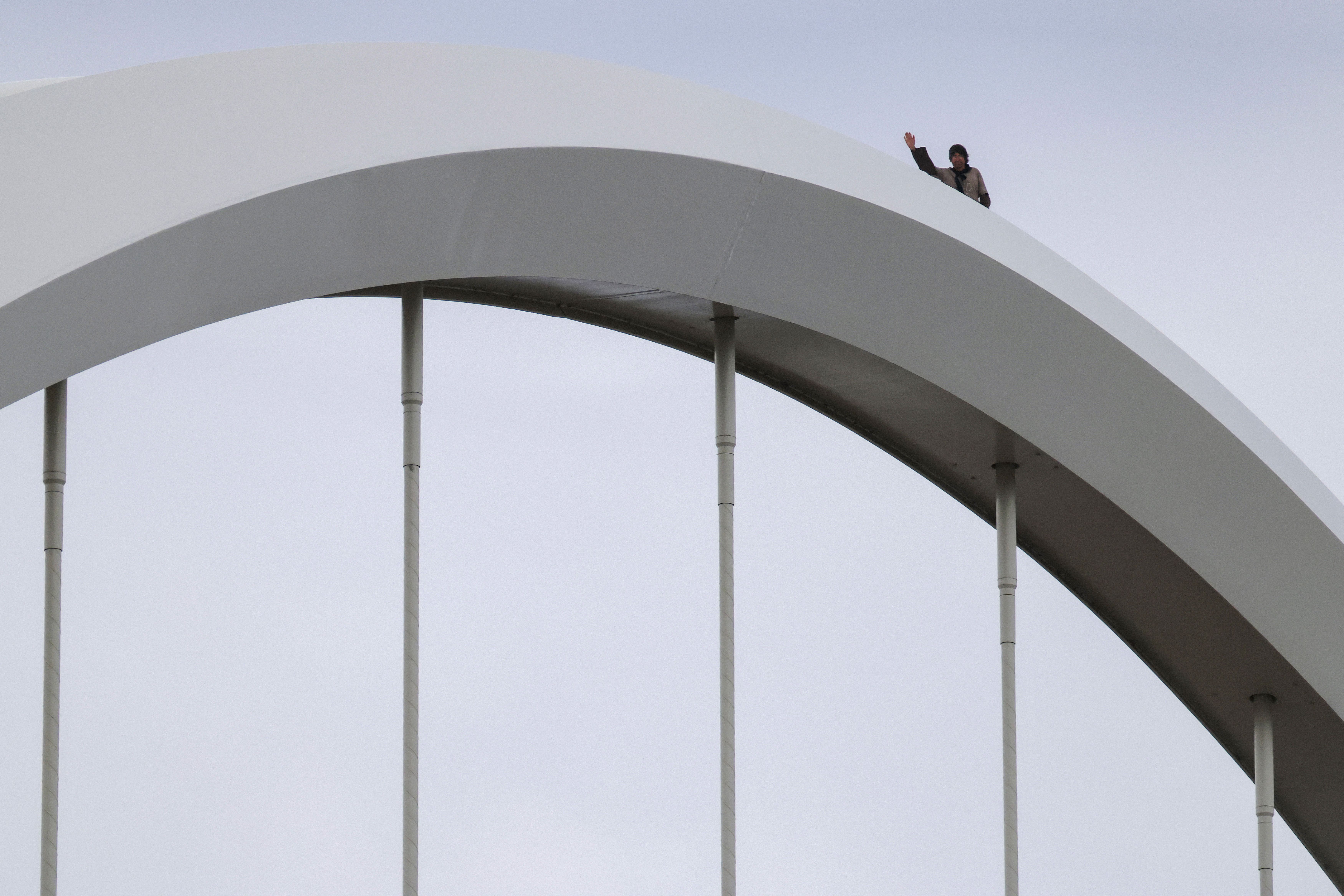 Person stands atop a large white curved arch, waving with one hand; the structure's arcs rise above vertical columns, set against a pale blue sky.