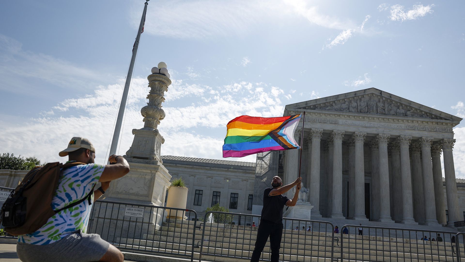 A person waves a pride flag outside of the Supreme Court while another person takes a photo.