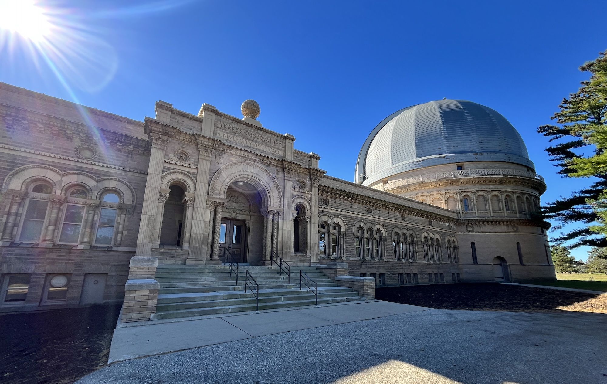 Bright day at Yerkes Observatory: an ornate stone building with arches and columns, and a large silver dome on the right. Clear blue sky, sun flare, and trees on the far right.