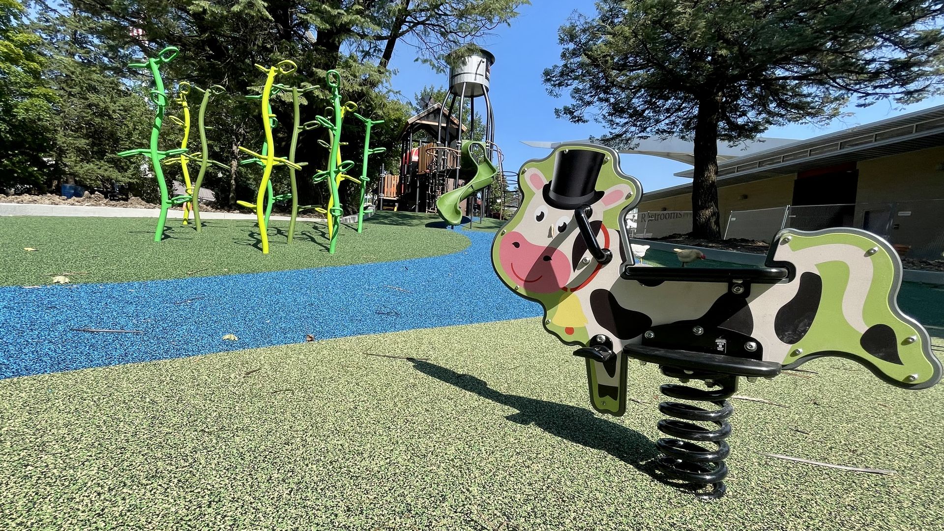 Colorful children's playground featuring a green and pink cow spring rider with a top hat, green climbing structures, a blue pathway, and a large play tower under a clear blue sky.