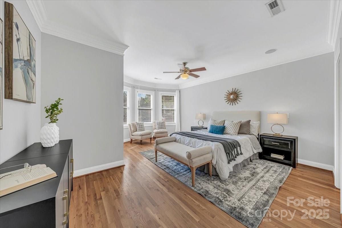 Bright bedroom with wood floors, gray walls, a bed with white and blue pillows, two nightstands with lamps, a bench, two chairs by bay windows, and a ceiling fan.