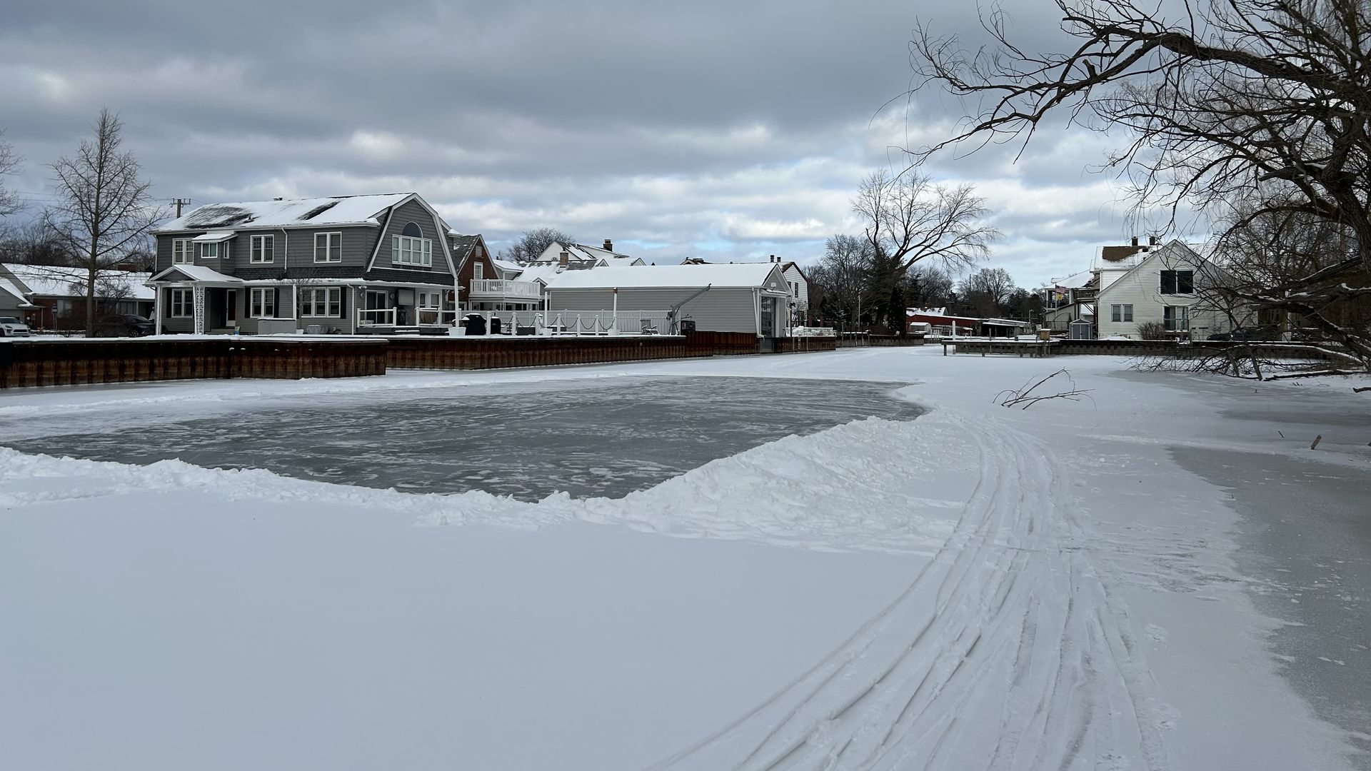 An ice rink has been made in the center of the snow that covers the canal. On either side of the canal, houses and metal seawalls.