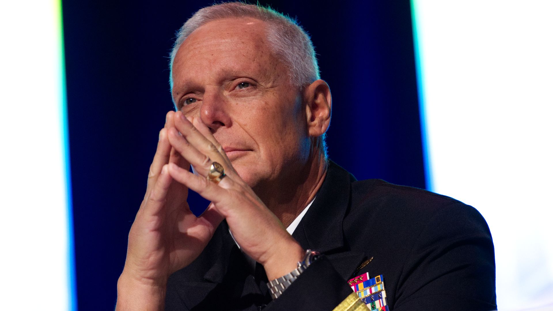 A man with short white hair, wearing a military uniform, peers over his hands while sitting behind a desk. He's at a professional event.