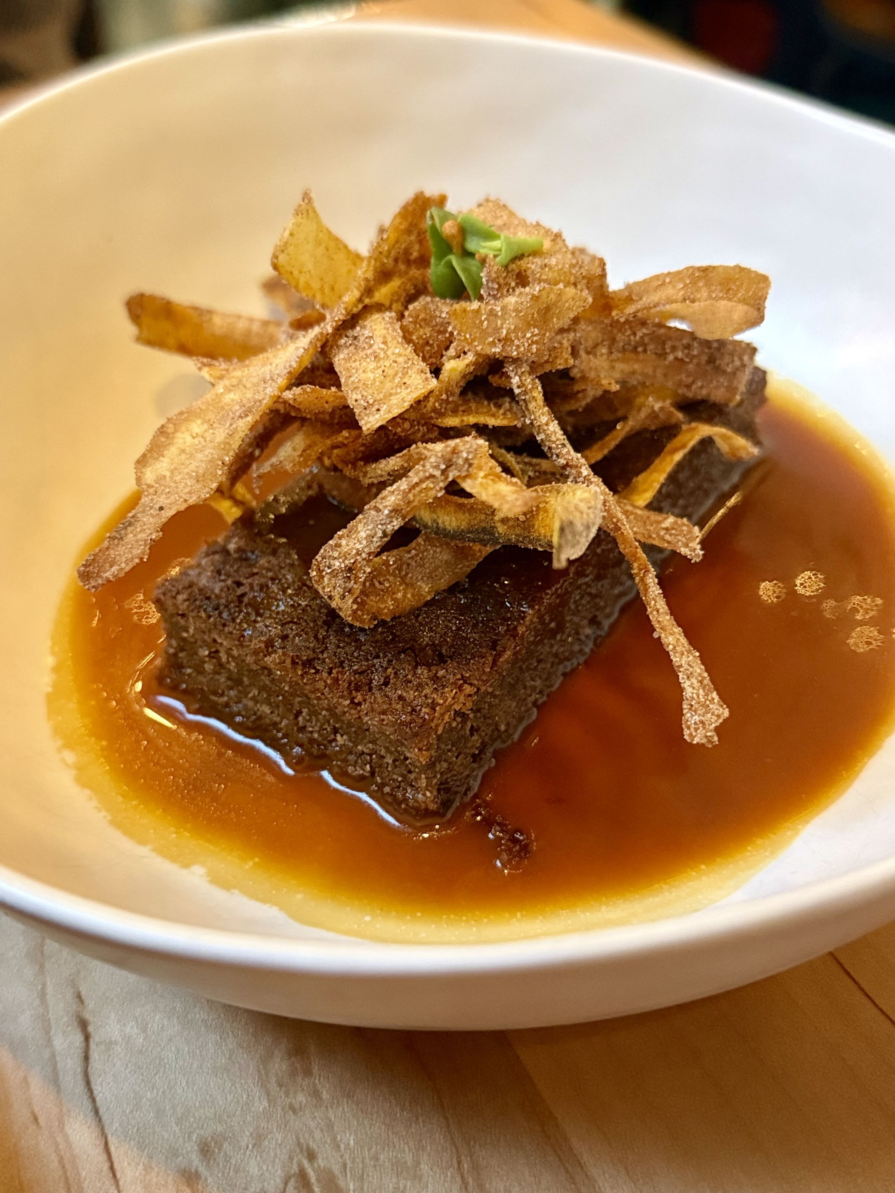 A white bowl holds braised meat in brown sauce topped with crispy thin strips (potato or tortilla) and a small green herb, adding a crunchy garnish over a rich, caramel-colored sauce.