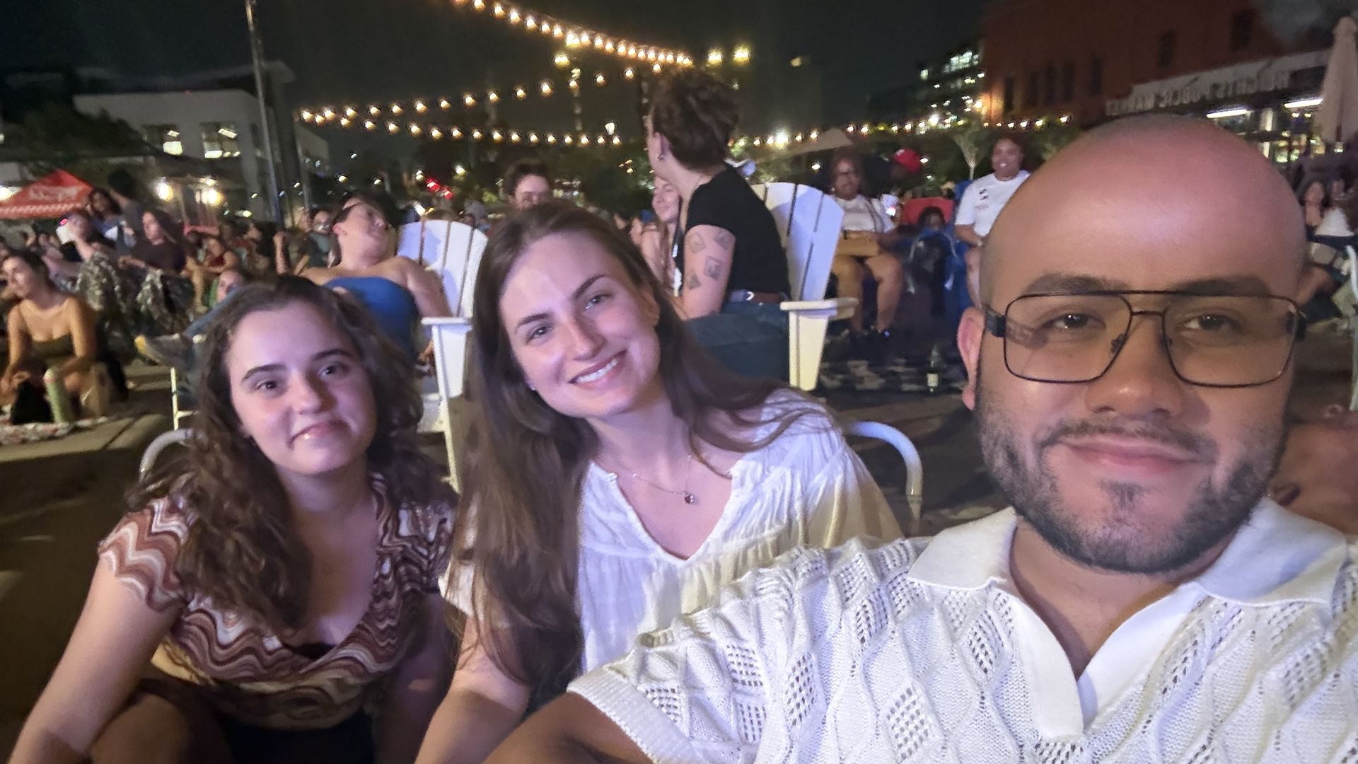 Selfie at a nighttime outdoor event with string lights. Three friends in the foreground: center woman smiling, left woman, right man wearing glasses; crowd seated behind.
