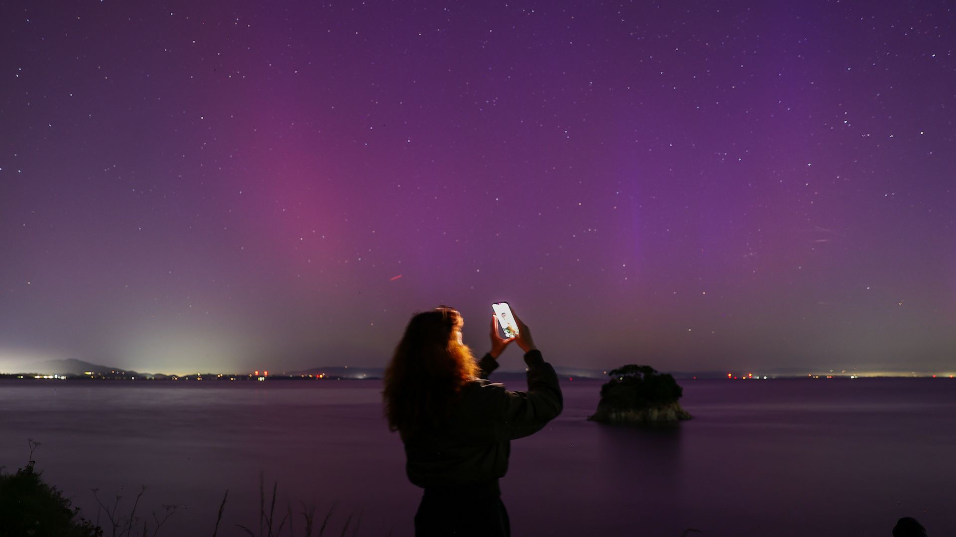 Woman holds smartphone to take photos of aurora and is standing in front of a colorful sky with pink, purple and green shades in sky
