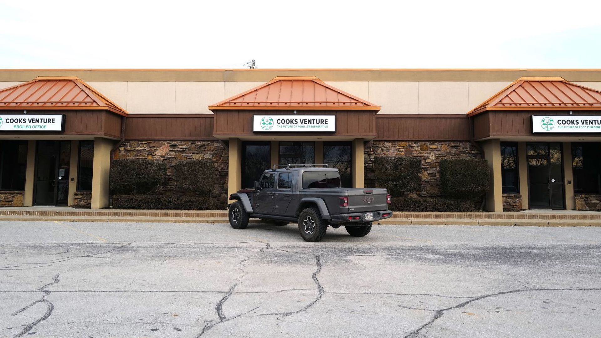 A photo of a building with three signs for Cooks Venture and a single four-door Jeep parked in the parking lot.