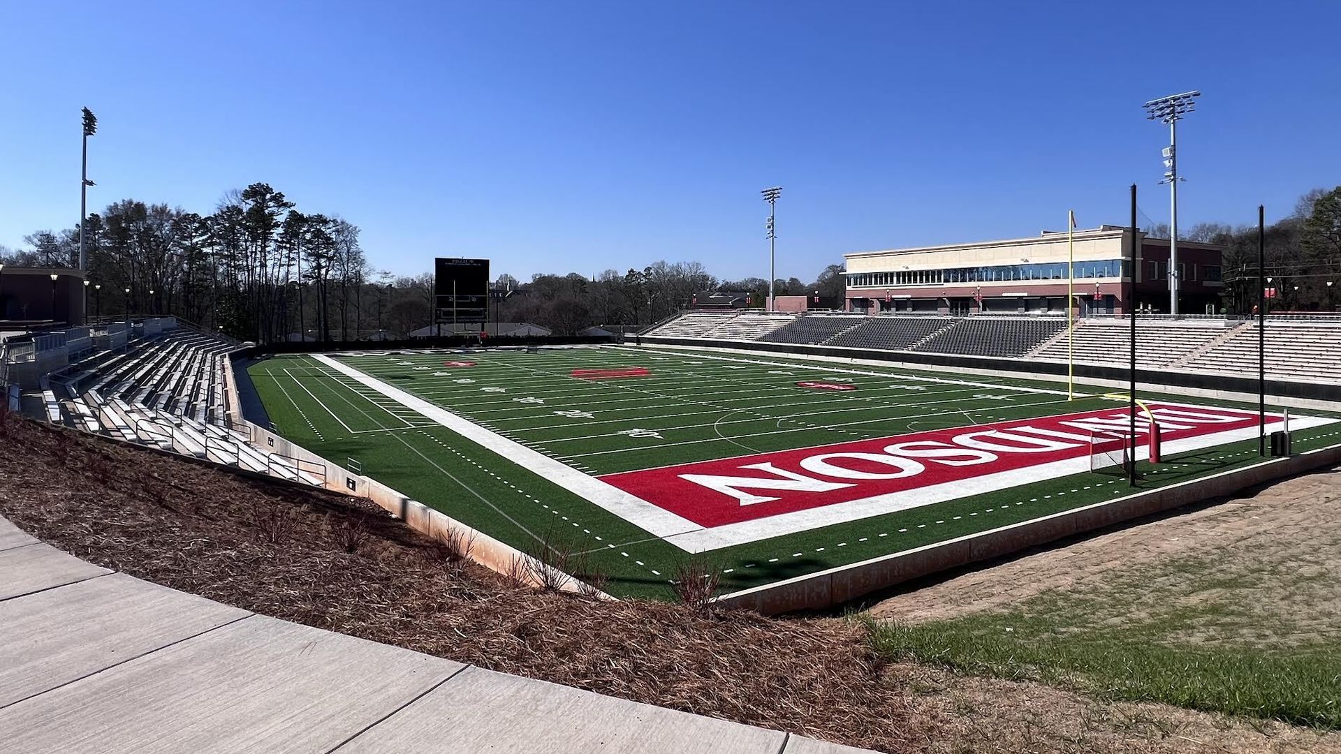 Empty football stadium with green field, white yard lines, red end zone marked "MADISON", gray bleachers, tall light poles, clear blue sky, and trees in the background.