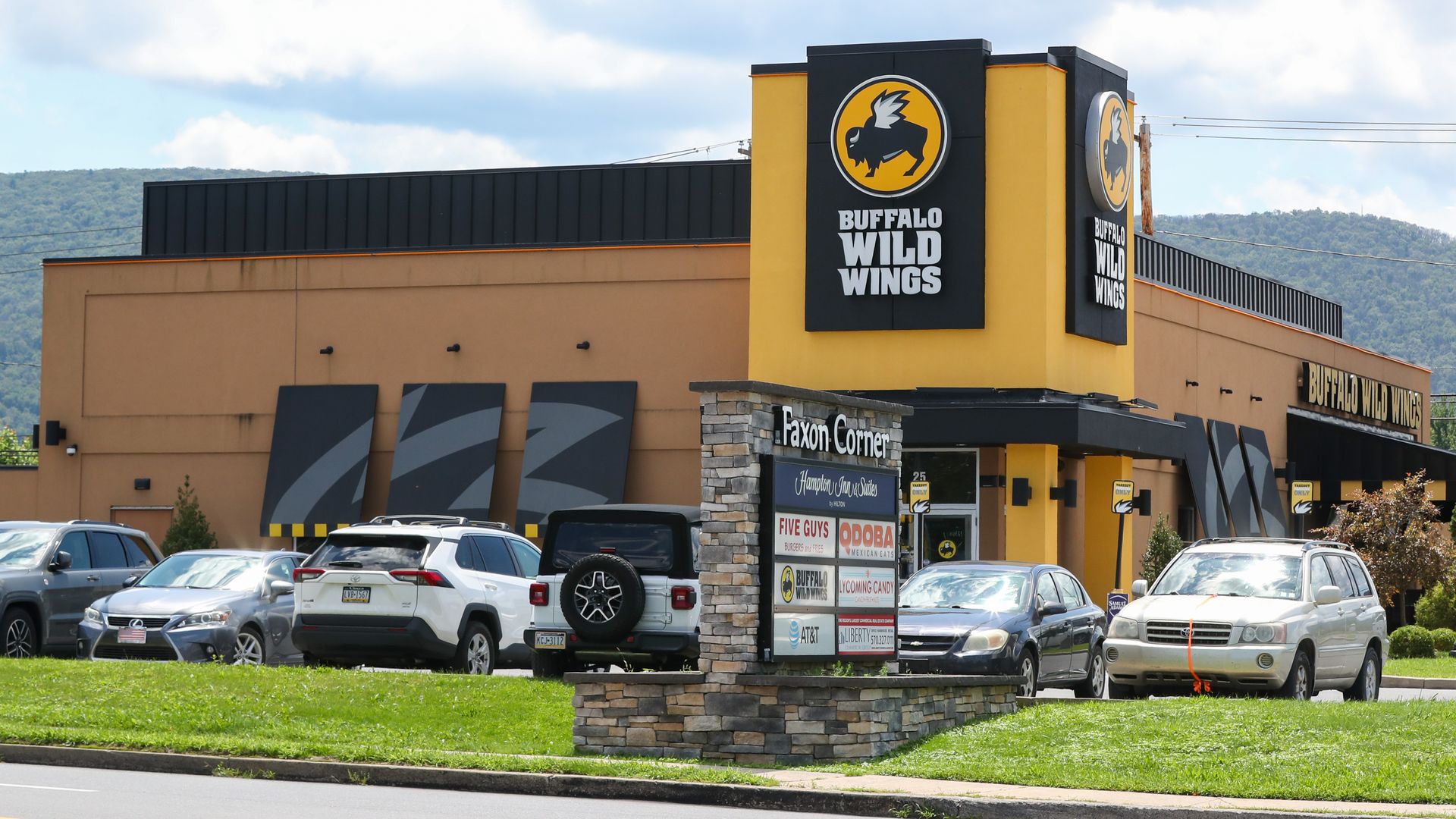 Buffalo Wild Wings restaurant with yellow and black branding, parked cars, and a sign for Faxon Corner showing multiple business logos including Five Guys and Qdoba under a partly cloudy sky.