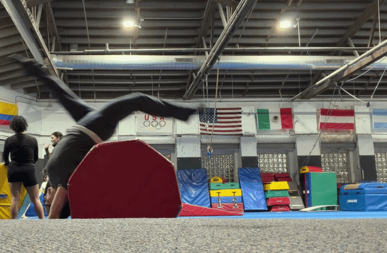 Indoor gymnastics gym with a motion-blurred gymnast flipping over a red padded mat; background stacks of blue and yellow mats, and flags (USA, Mexico, others) under a metal ceiling.