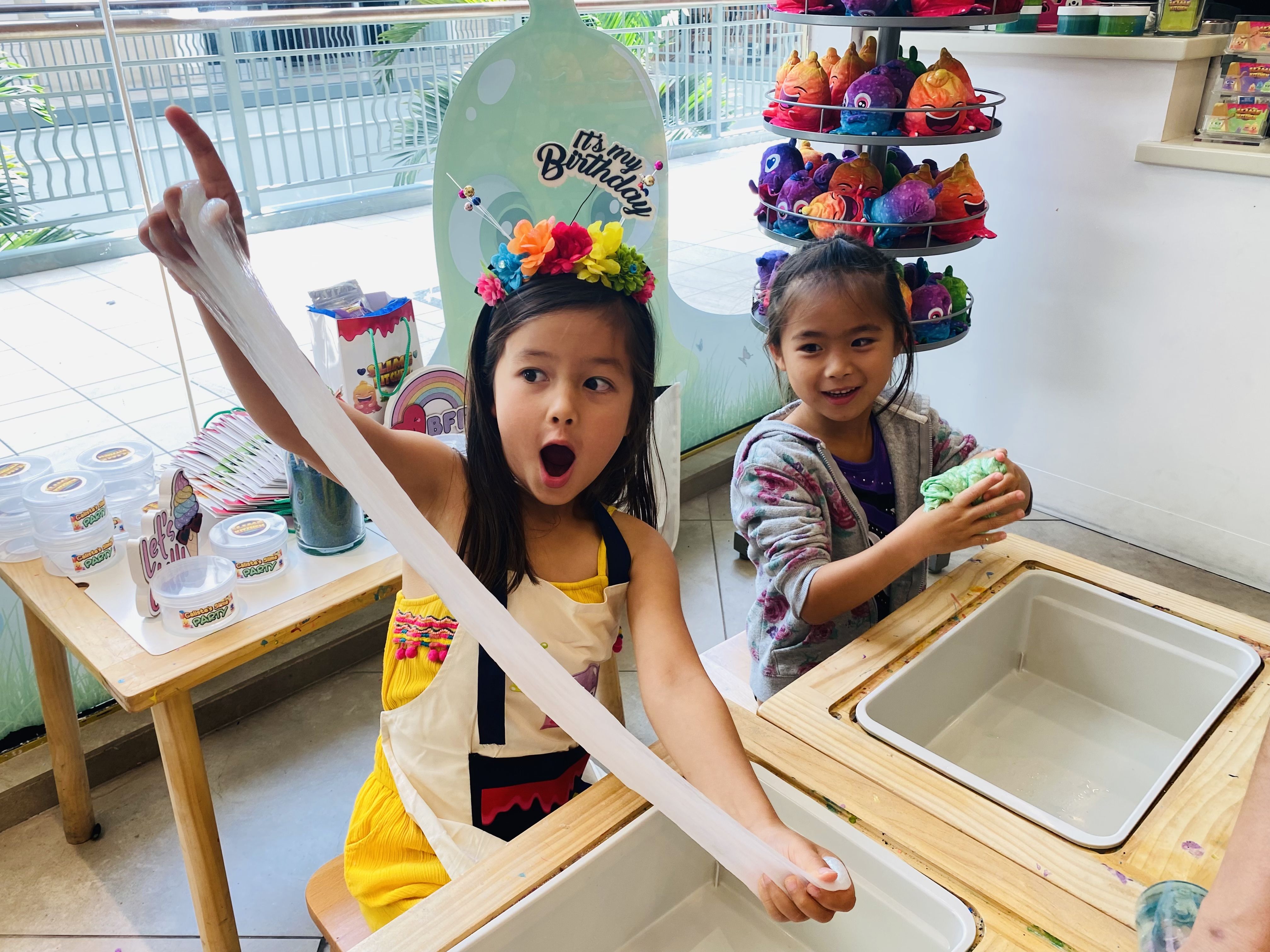 Two girls playing with slime at a wooden table; one in a yellow dress and flower crown stretches white slime, the other in a hoodie holds green slime, colorful plush toys in background.