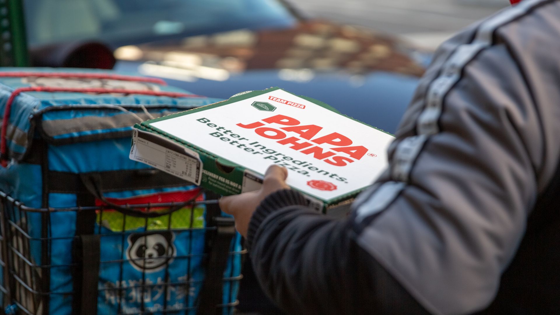Person holding a Papa John's pizza box near a bicycle basket with a blue delivery bag that has a panda logo, with a blurred car in the background.