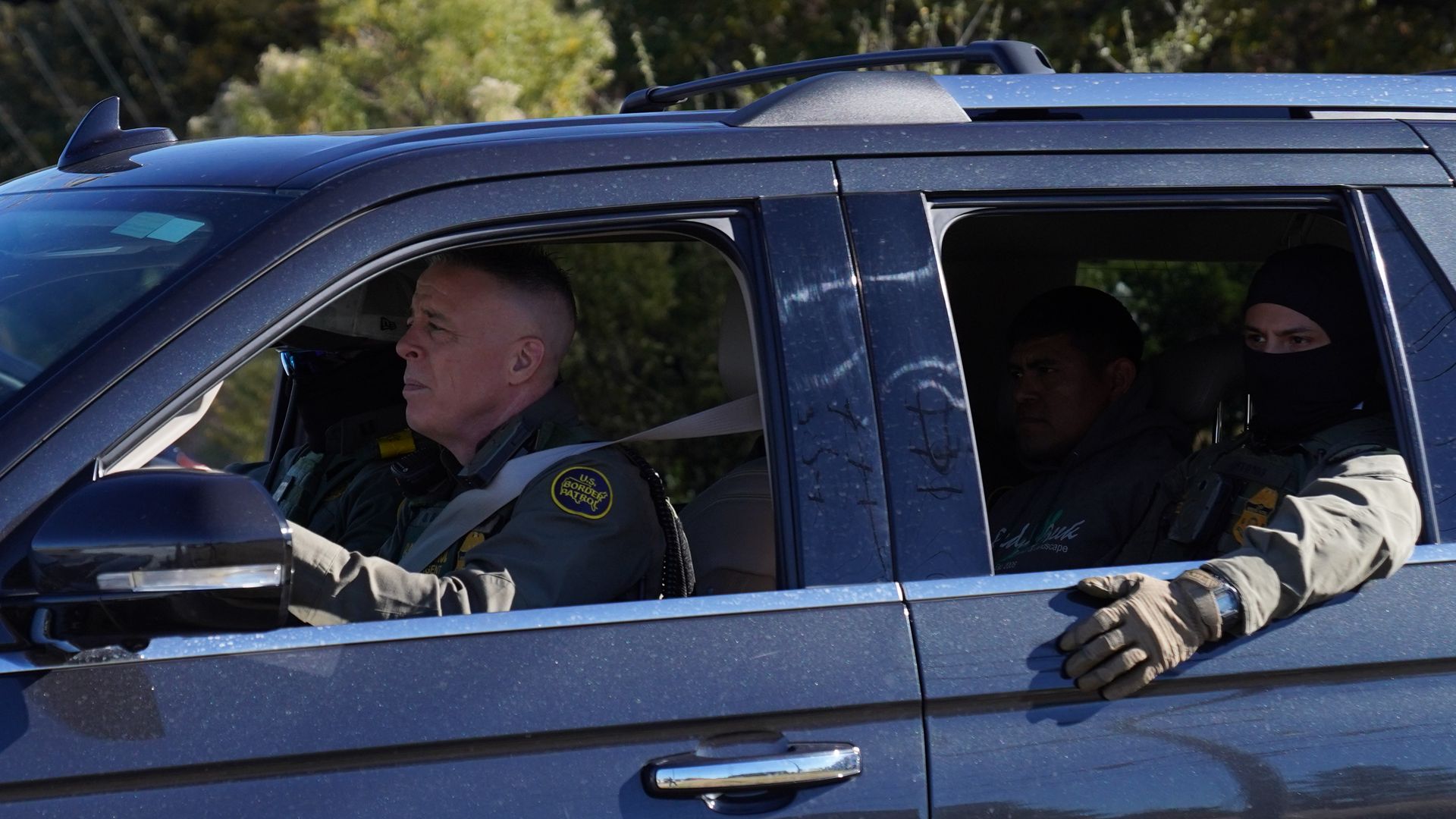 Blue SUV with U.S. Border Patrol agents, one masked with gloves, driving with a detained person sitting in the back seat, trees in the background.