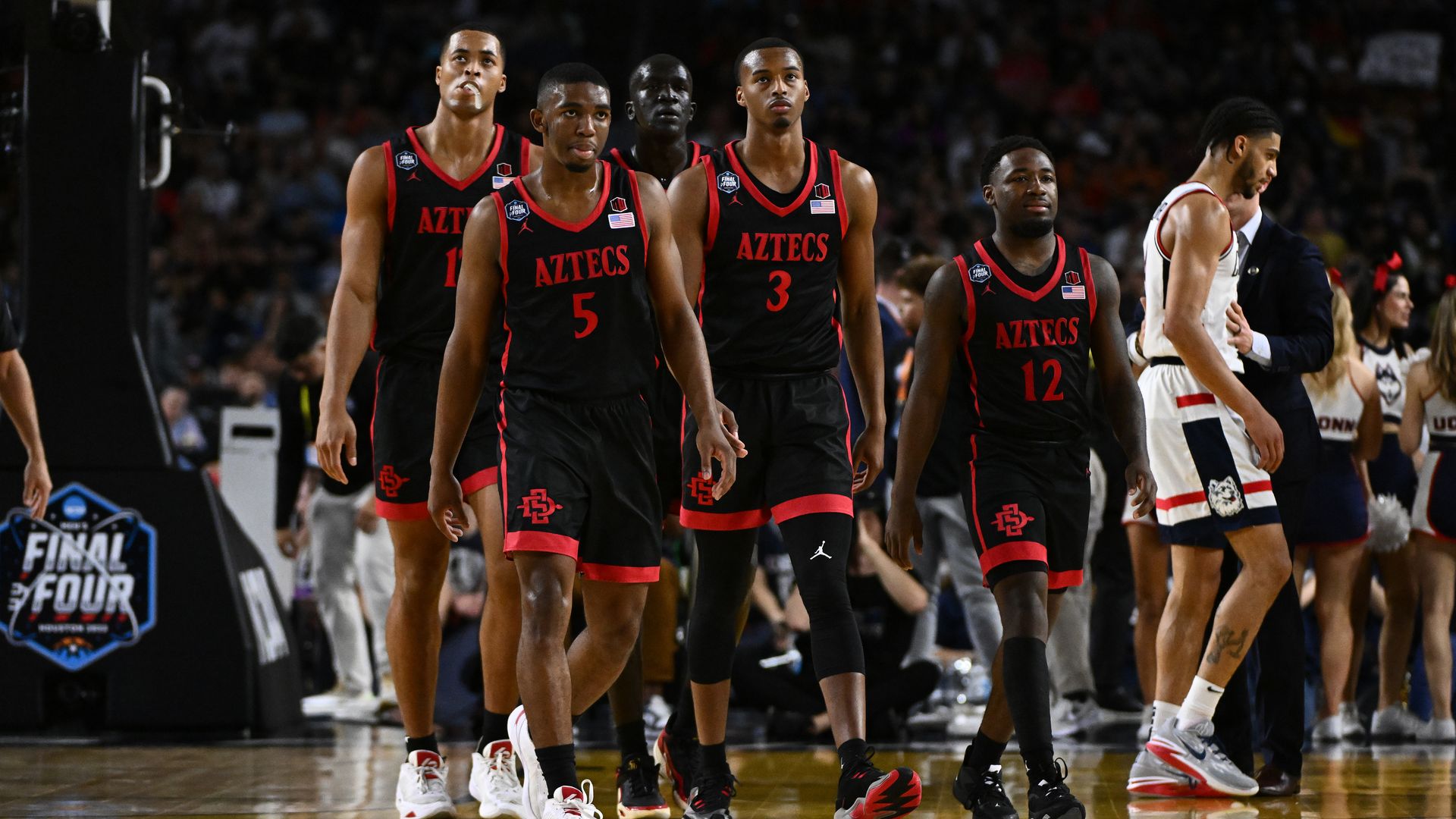 Five basketball players in black and red uniforms walk on the court together. 