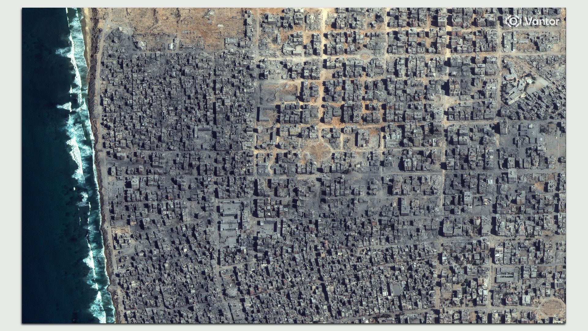 Satellite image showing a large area of destroyed urban buildings near a coastline with visible ocean waves.