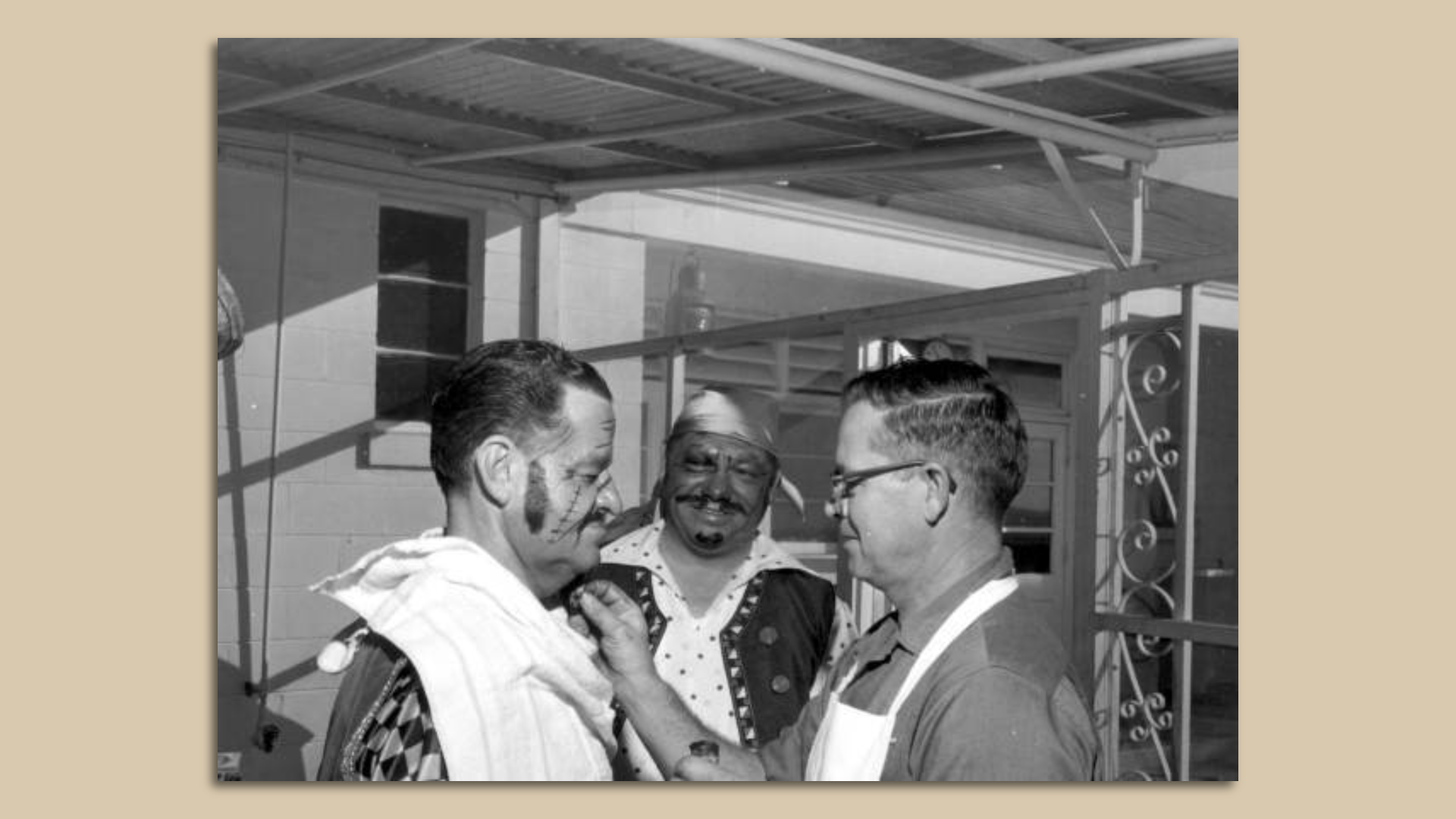 A man applies another man's pirate makeup while a third man watches on, smiling.