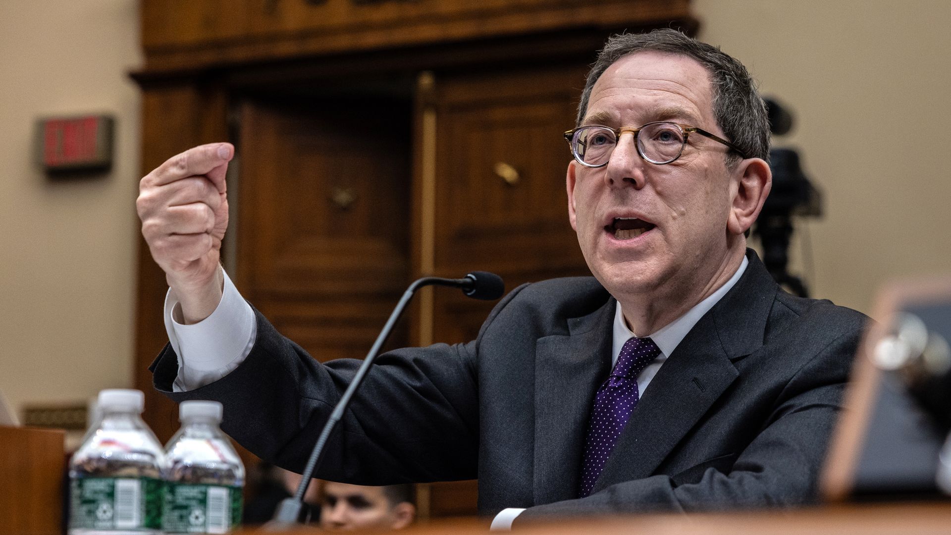 Photo of a man gesturing behind a microphone in front of a desk. 