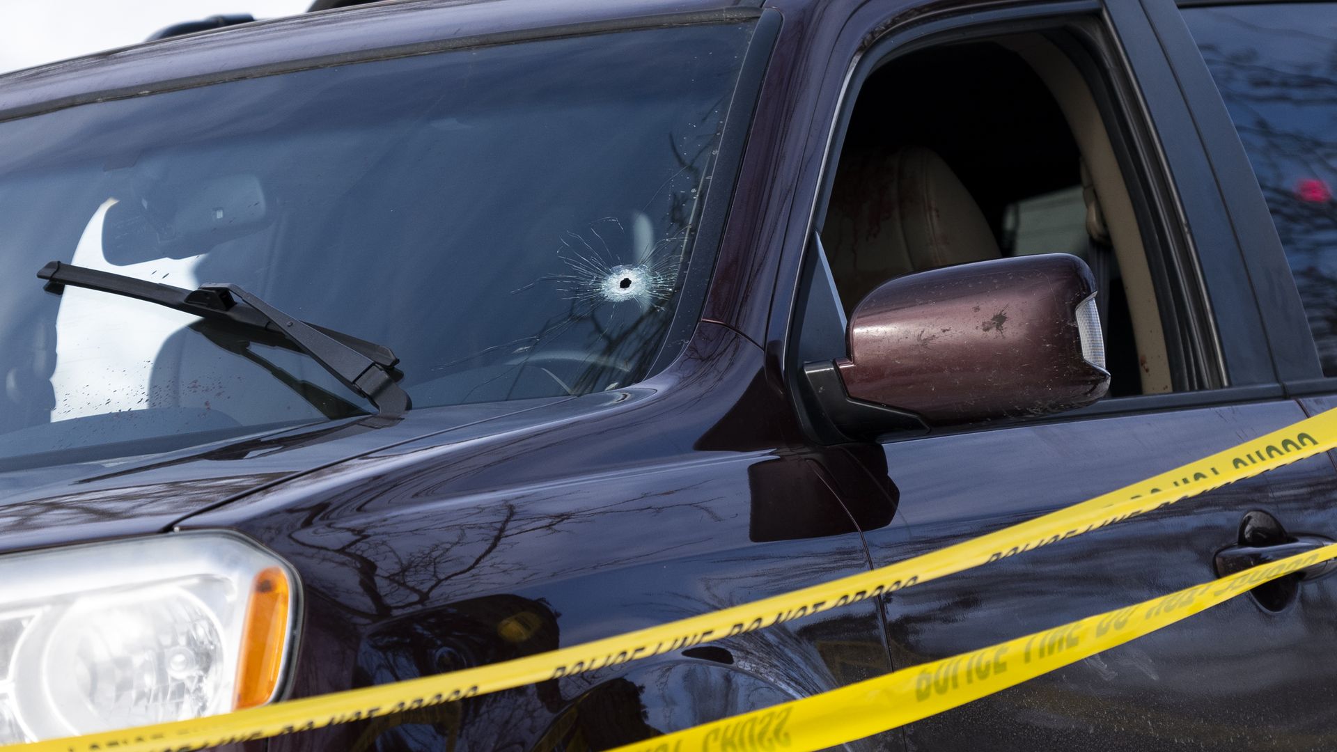 Dark SUV with a shattered windshield and a visible bullet hole near the driver's side; yellow crime tape stretches across the foreground, while a blurred figure wearing a helmet stands behind.